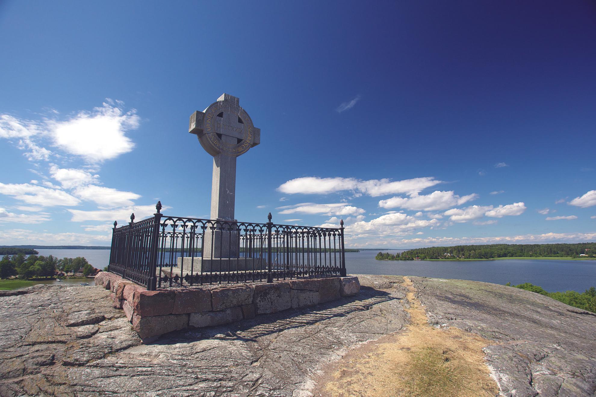 Das Kreuz von Ansgar steht auf einer kahlen Klippe. Man kann den See Mälaren, kleine Inseln und einen blauen Himmel im Hintergrund sehen.