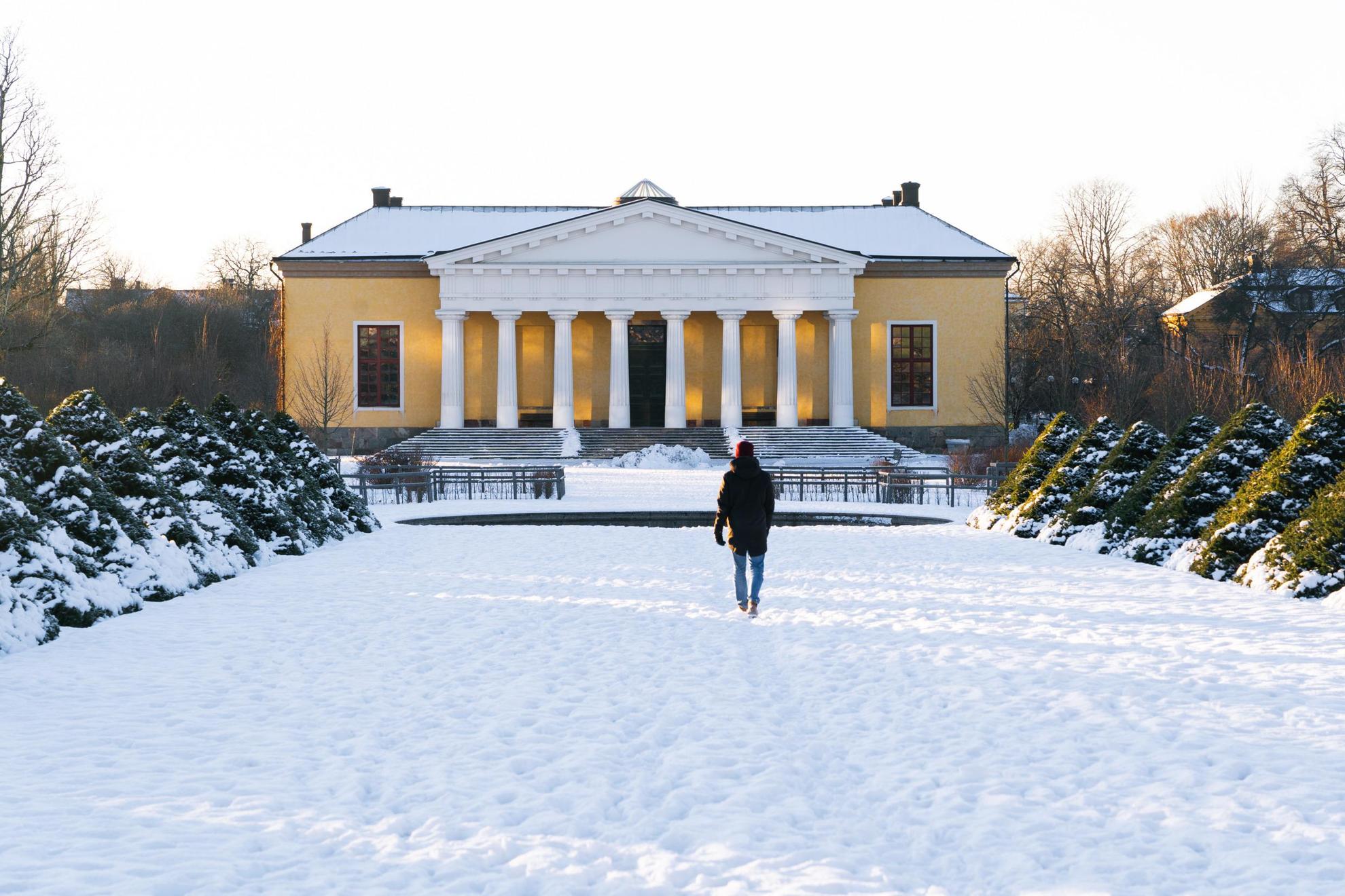 Eine Person, die im Winter auf ein gelbes Haus im botanischen Garten der Universität Uppsala zugeht.