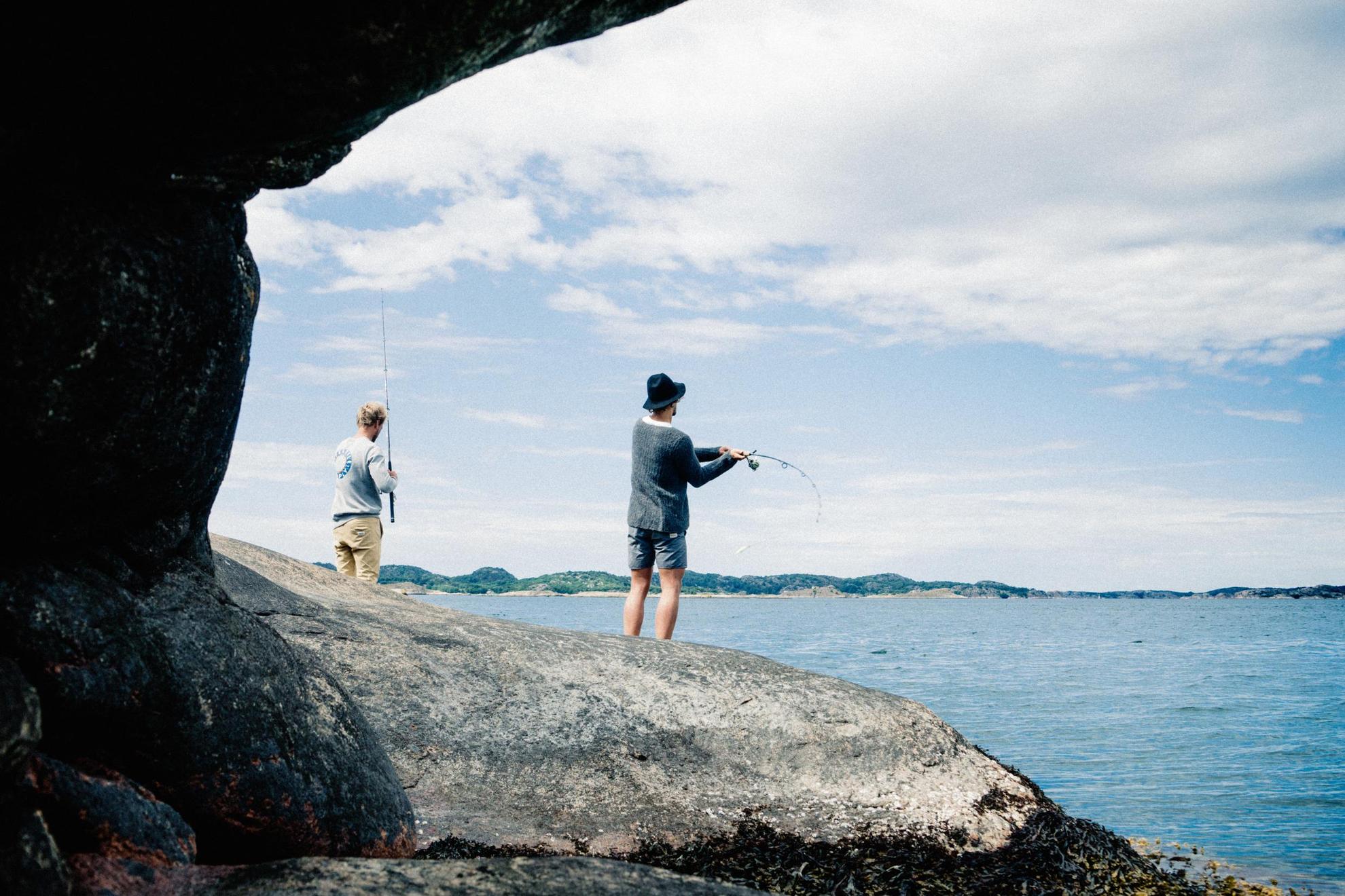 Zwei Personen stehen an der Küste  und Angeln. Im Vordergrund sind Felsen und am Horizont das Ufer einer Insel im Schärengarten.