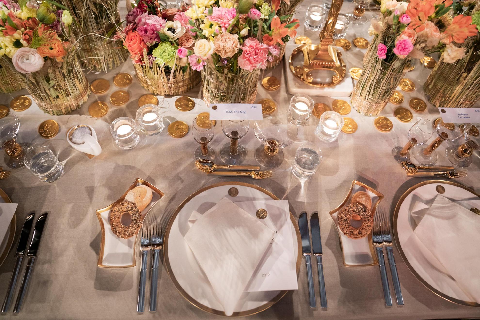 Ein Geschirr in Weiß mit goldenen Details wird auf einem Tisch mit einer weißen Tischdecke gedeckt. In der Mitte des Tisches stehen bunte Blumen in Vasen.