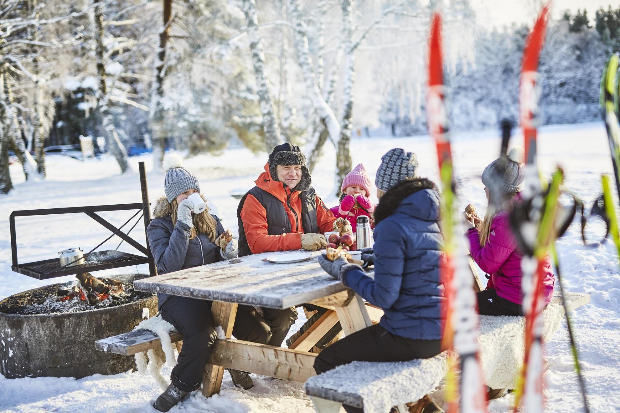 Eine fünfköpfige Familie sitzt draußen an einem Tisch, isst Zimtschnecken und trinkt Kaffee. Im Vordergrund des Bildes stehen Skier.