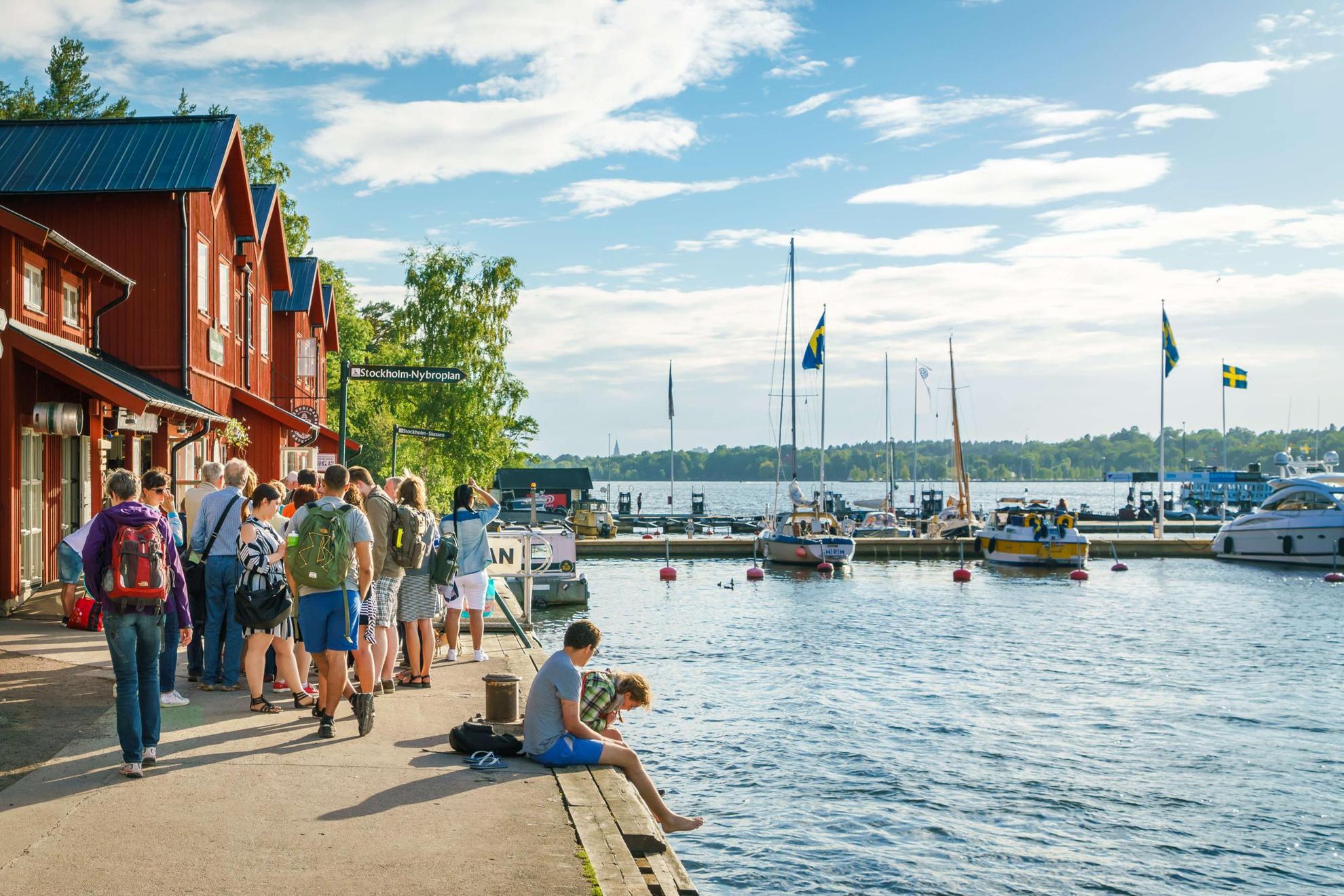 Menschen sitzen an einem Hafen in Schweden. Das Sonnenlicht fällt auf das Wasser. Es stehen Boote im Hafen und am Ufer stehen rote Häuser.