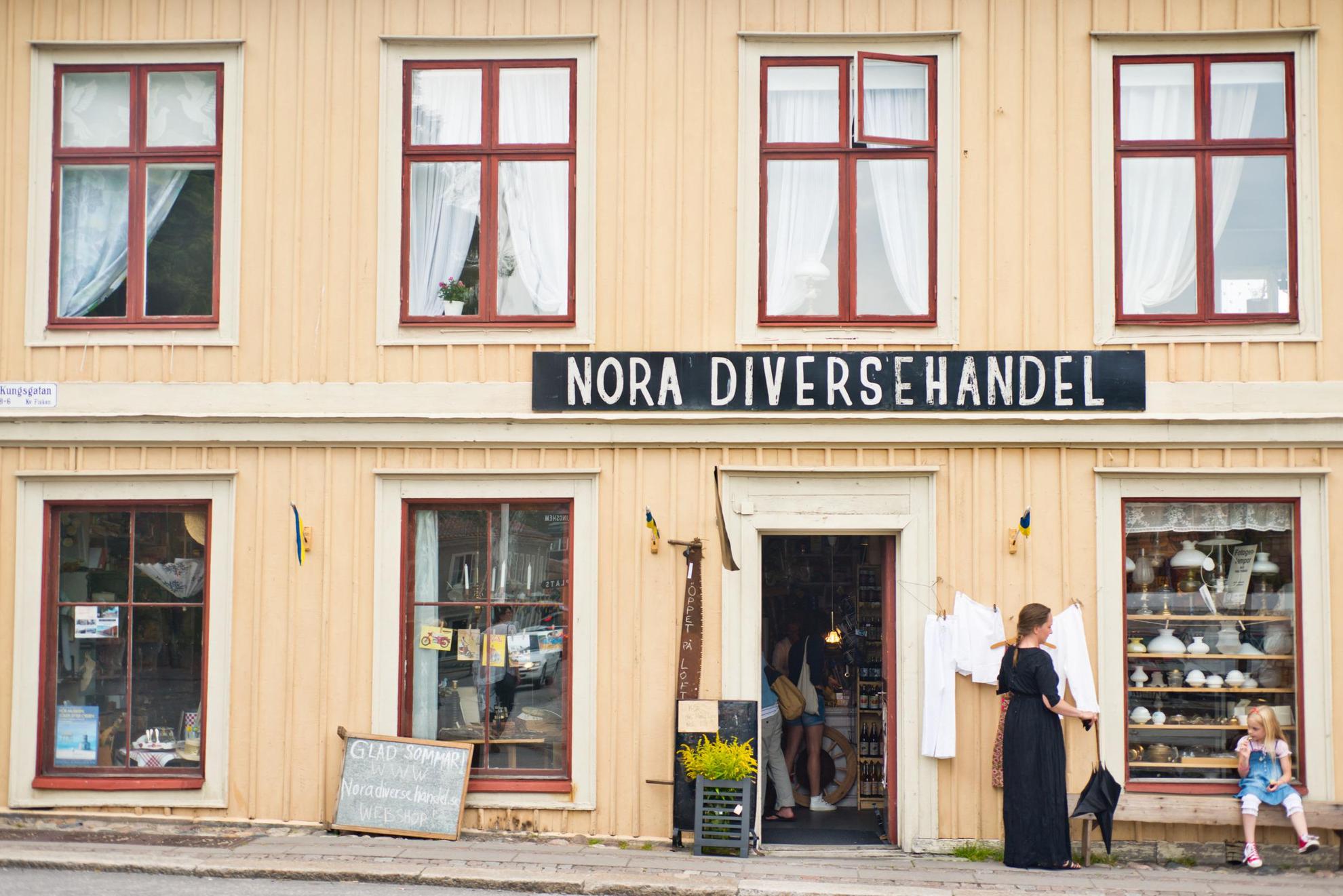 Ein kleiner Laden in einem gelben Holzhaus an einer Straße in einer Altstadt. Auf einem Schild über dem Eingang steht Nora Diversehandel. Eine Frau steht und ein Mädchen sitzt auf einer Bank vor dem Haus.
