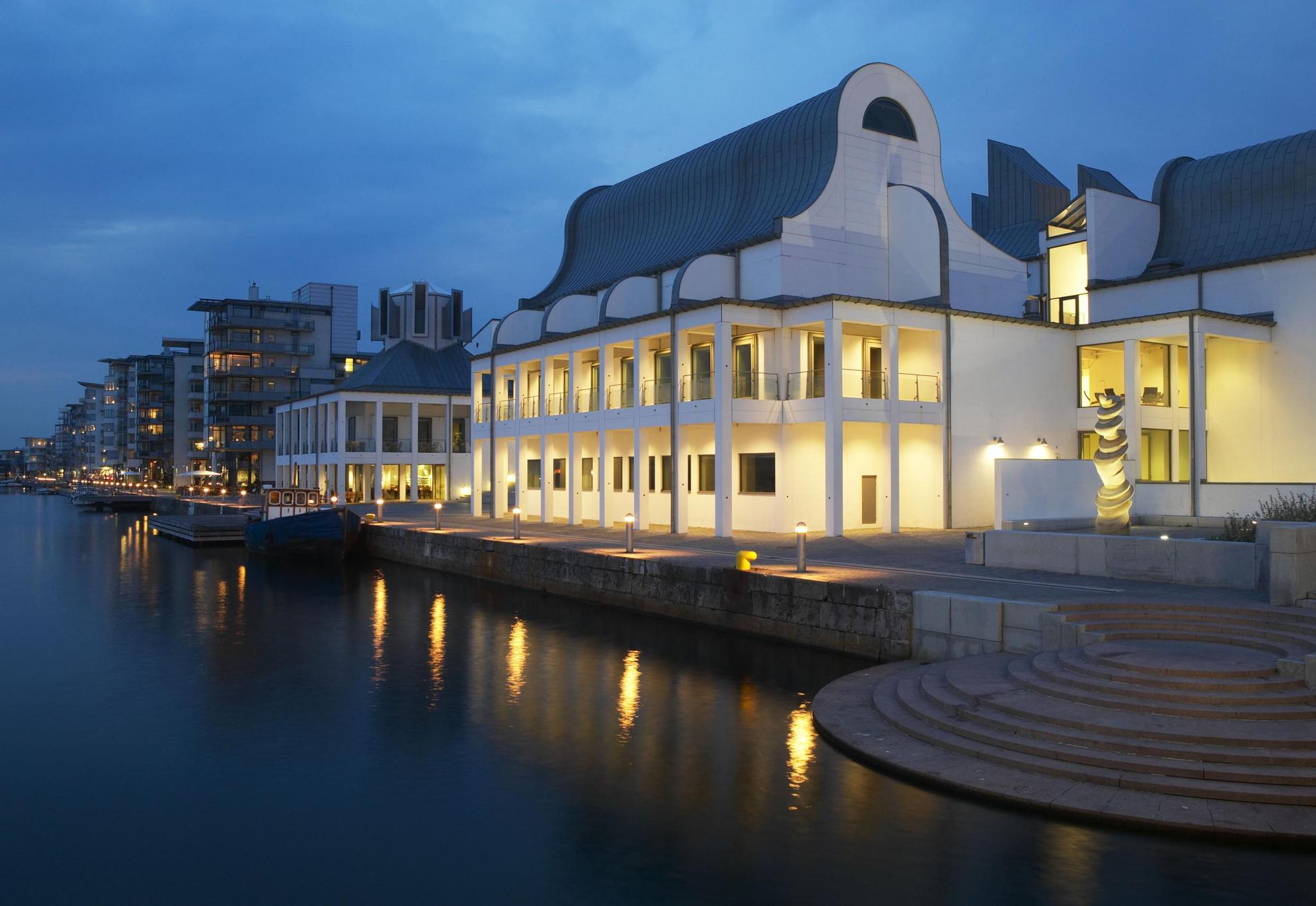 Dunkers Kulturzentrum im Abendlicht und mit Lampen an der Vorderseite, direkt am Meer an einem Jachthafen gelegen. Im Hintergrund siehst du Gebäude und Boote am Ufer.