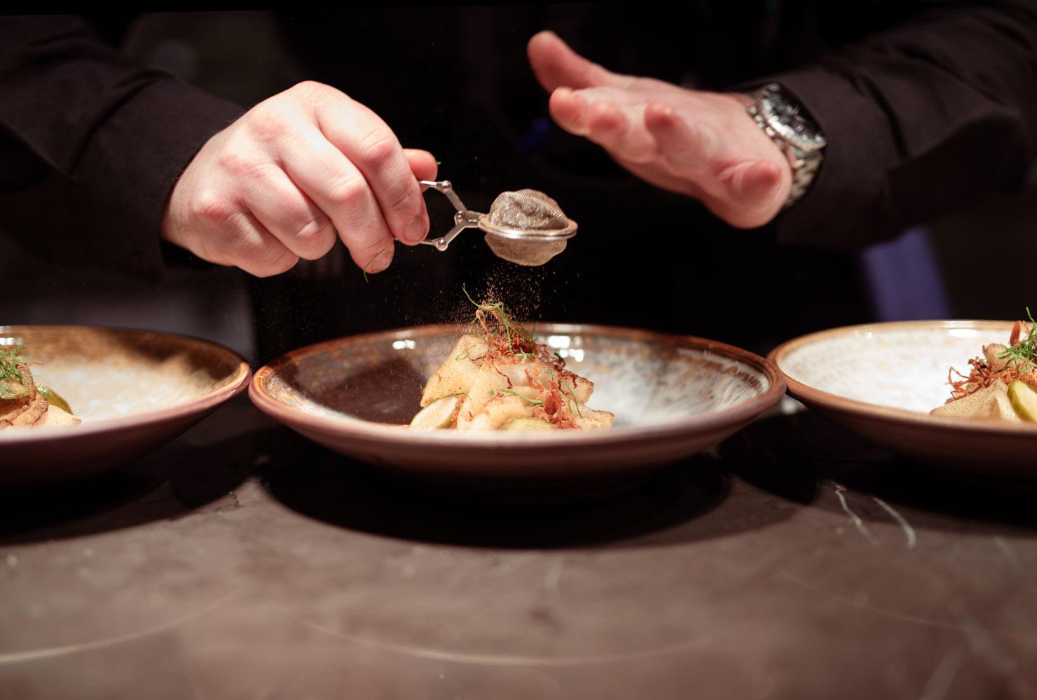 A close-up of two hands dusting a powder over a dish.