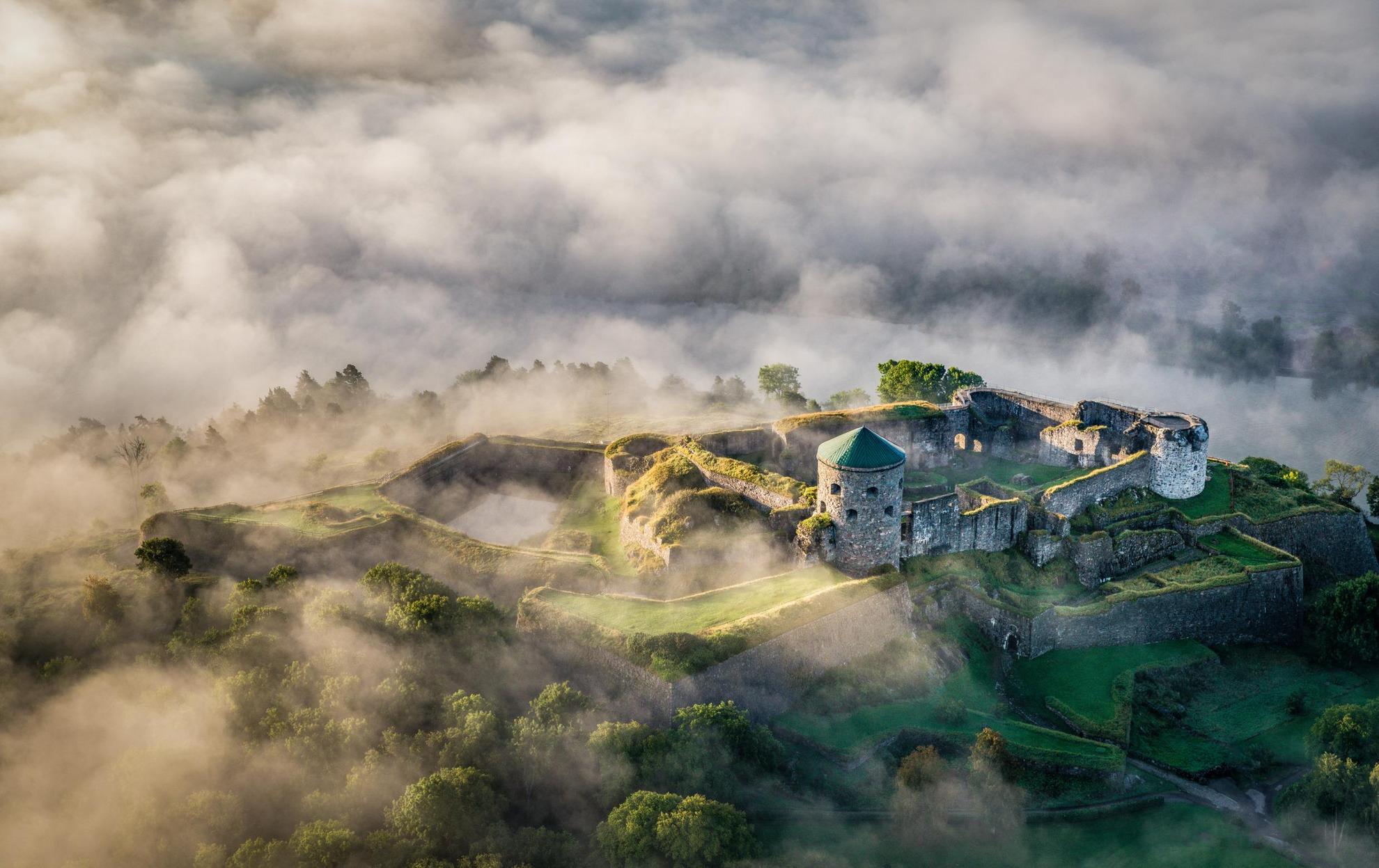 Luftbild auf einen Hügel, auf dem eine Festung mit Türmen und Mauern zu sehen ist. Wolken und Nebel ziehen über die Landschaft.