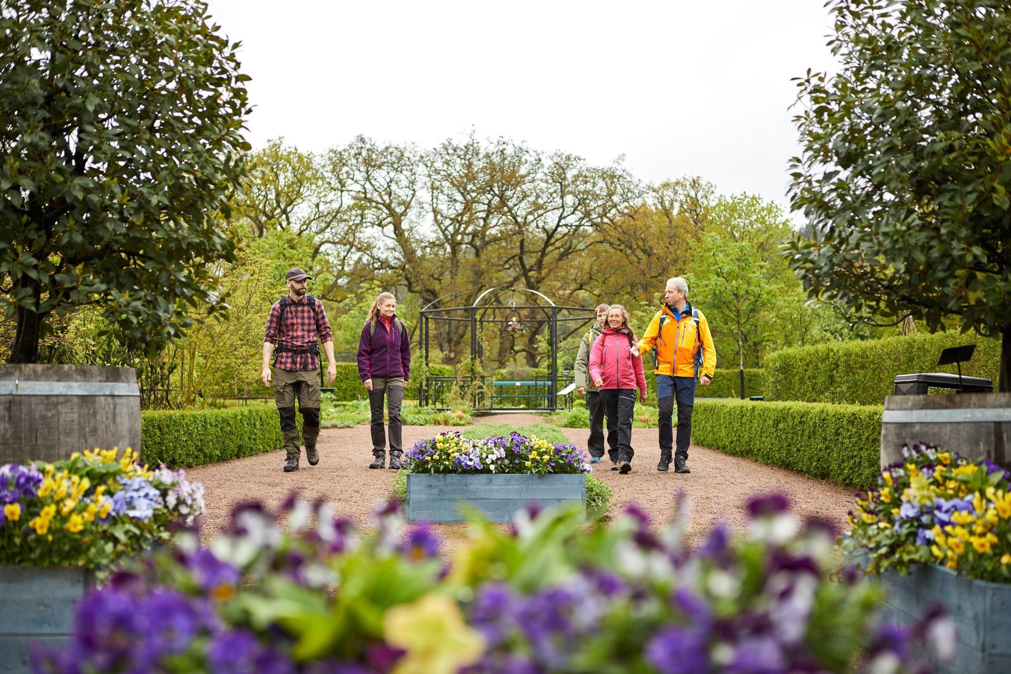Eine Gruppe von fünf Personen, die in einem wunderschön angelegten Garten spazieren gehen. Lila und gelbe Blüten am vorderen Bildrand. Im Hintergrund sieht man Bäume und Grün.