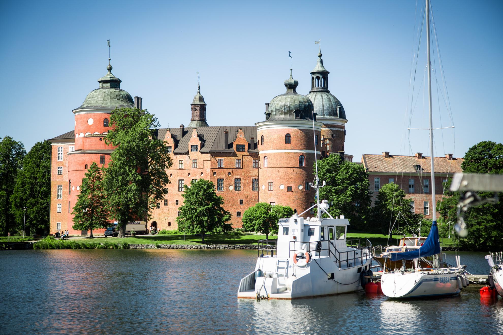 Das von Grün umgebene Schloss Gripsholm liegt auf der anderen Seite des Sees Mälaren. Die Sonne scheint und Boote liegen am Pier.