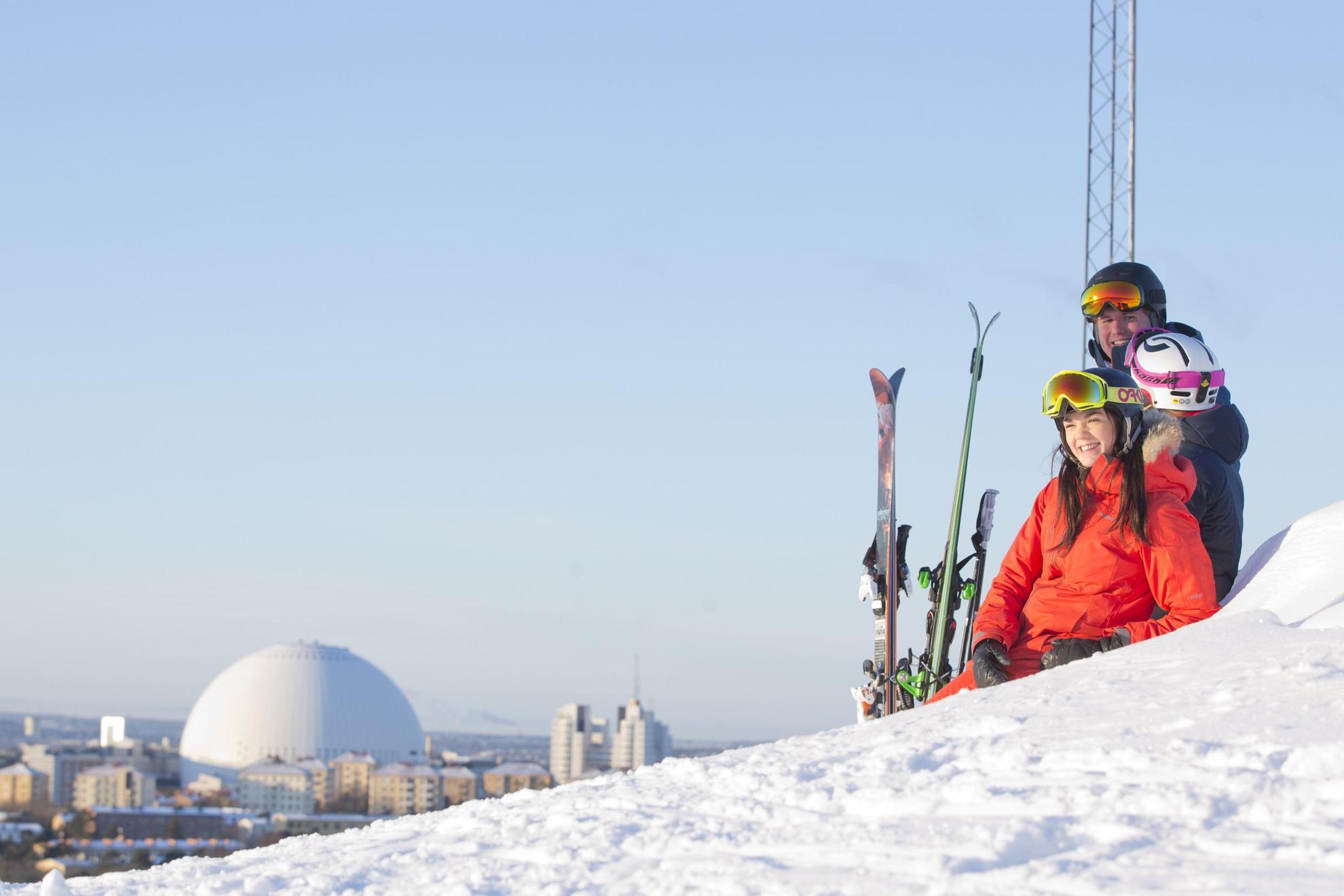 Drei Personen in Skiausrüstung entspannen sich im Schnee und genießen die Aussicht auf Stockholm.