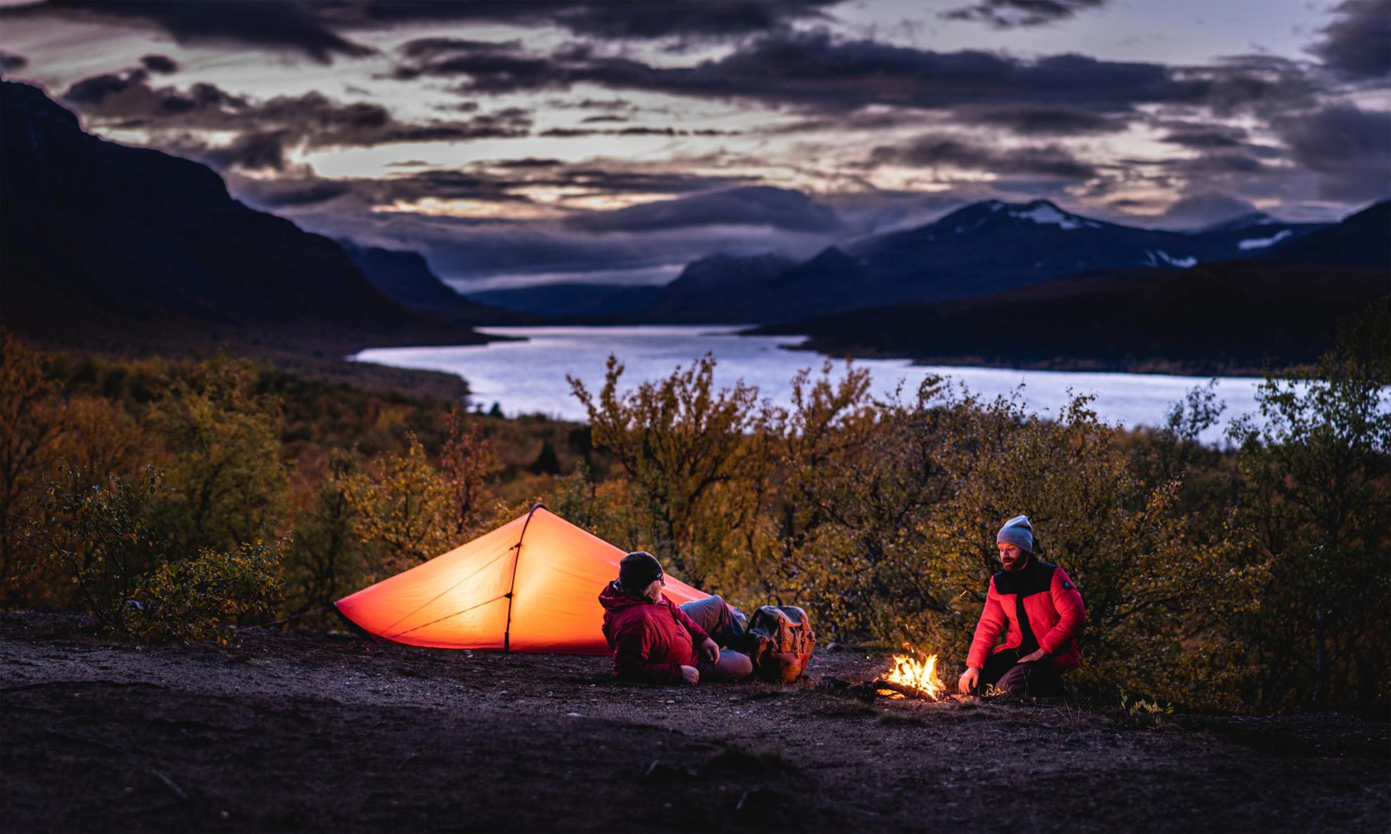 Zwei Personen sitzen neben einem Zelt und einem Lagerfeuer. Ein Fluss und Berge sind im Hintergrund.