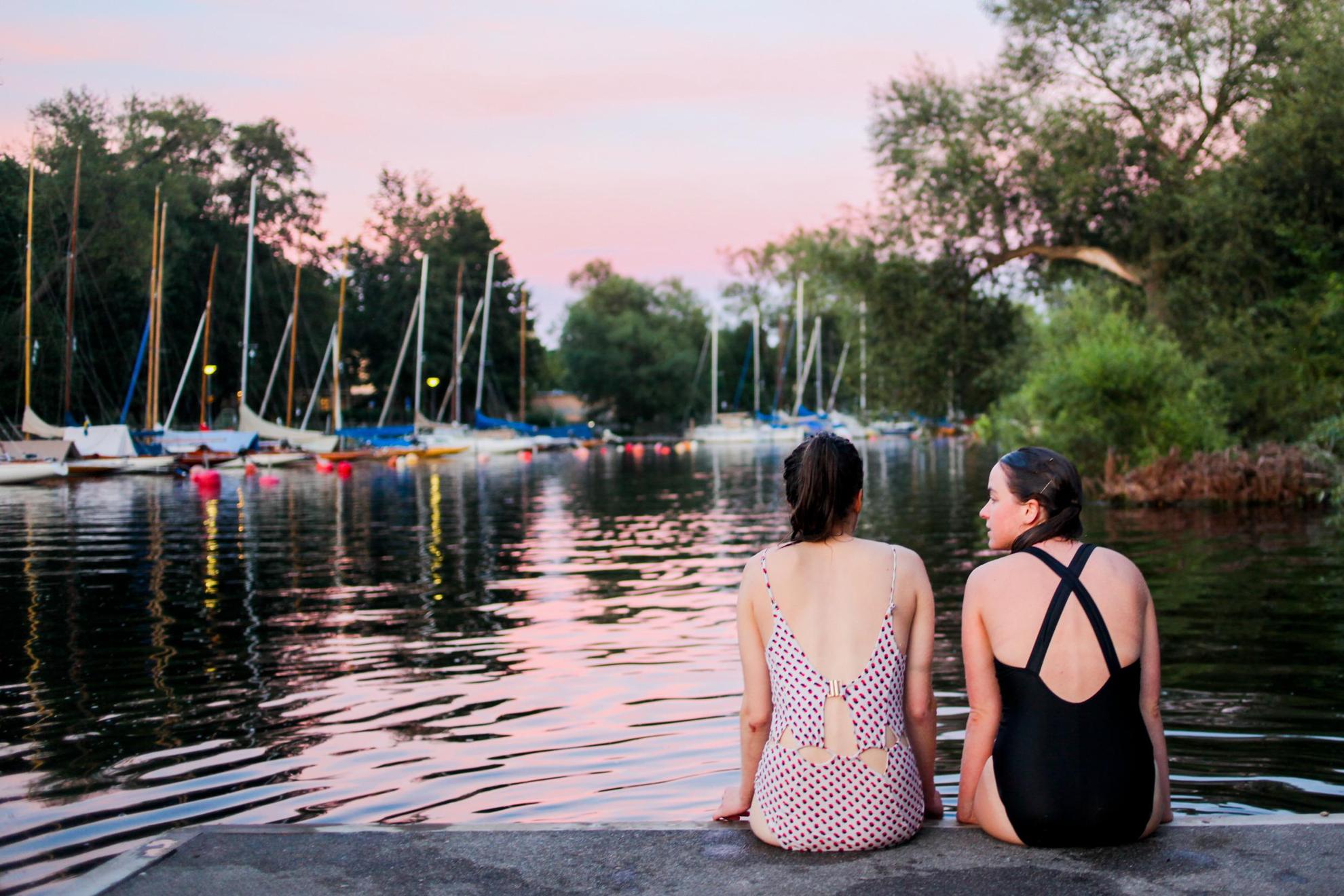 Two girls are sitting on a pier across from moored private boats.