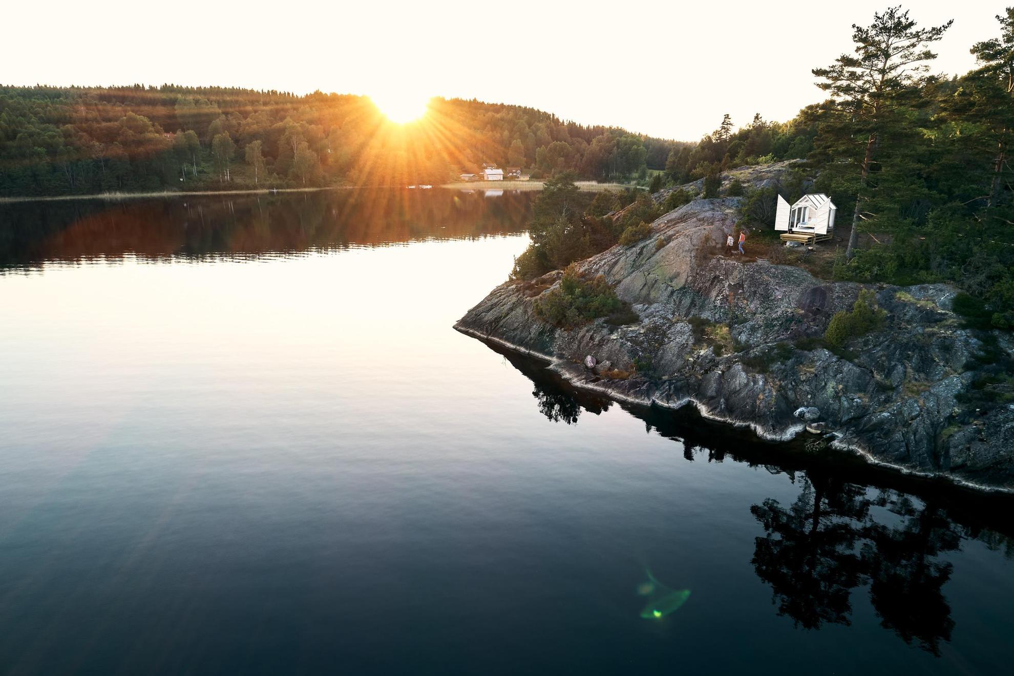 Blick auf eine Glashütte auf einer Klippe an einem See. Vor der Kabine laufen zwei Personen. Die Sonne geht hinter dem Wald unter.