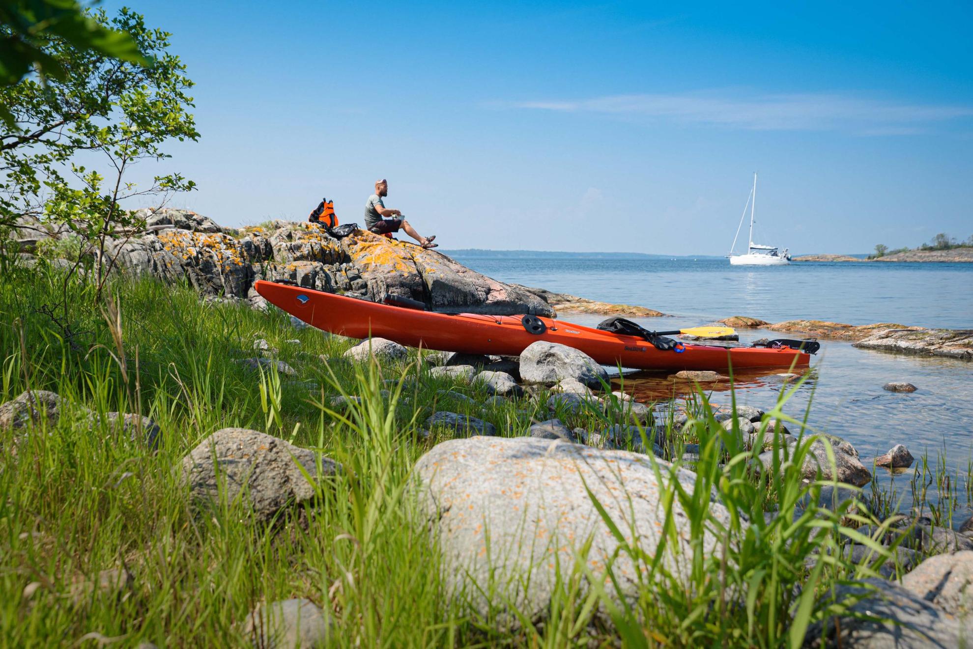 Ein orangefarbenes Kajak liegt am Wasser, während ein Mann auf den Klippen zu Mittag isst. Im Hintergrund ist ein weißes Segelboot zu sehen.