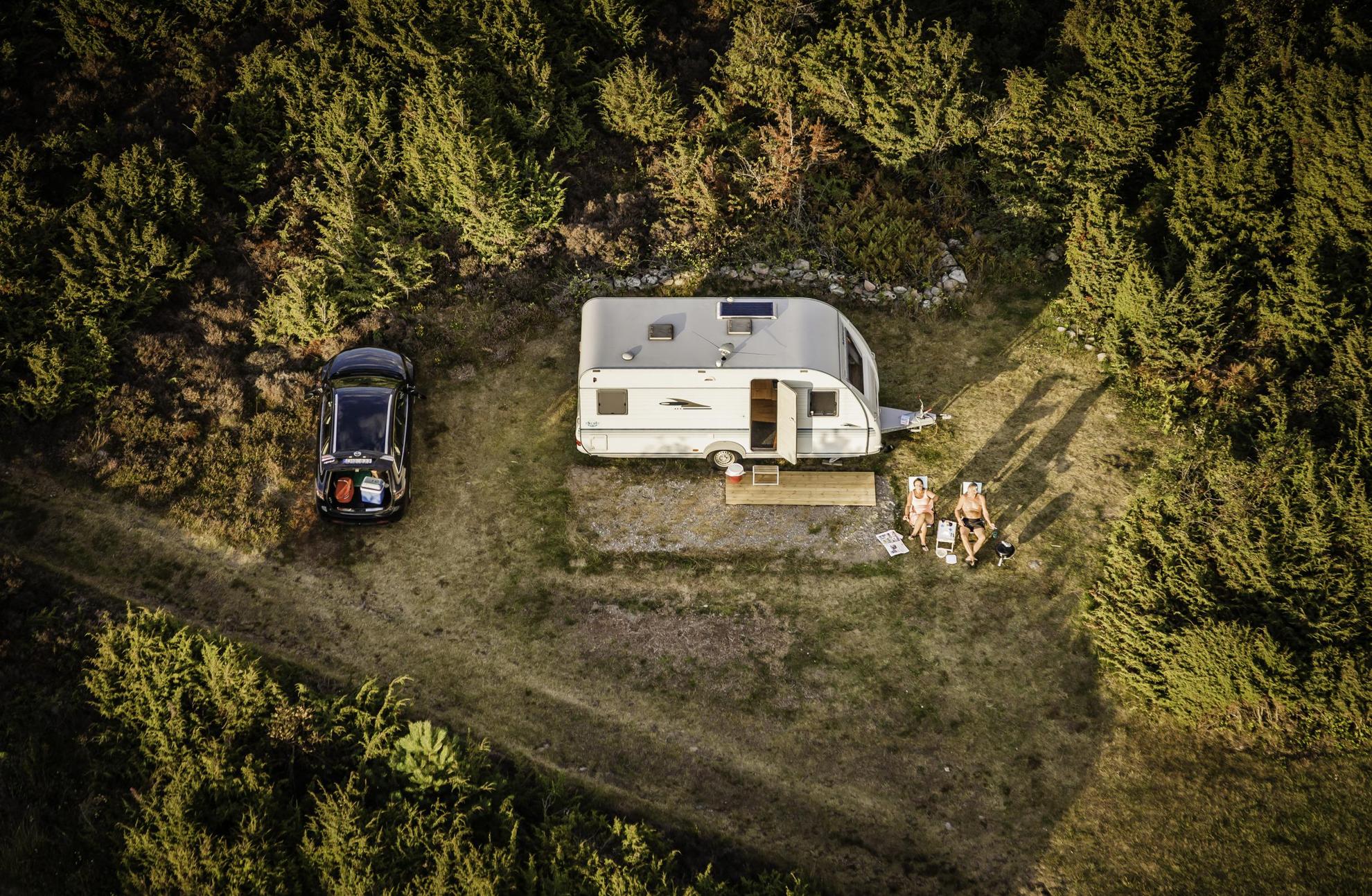 Zwei Personen Campen in Schweden. Ein Wohnwagen steht auf einem Wohnmobilstellplatz auf einem Campingplatz in Schwedens Wäldern. Zwei Personen sitzen in ihren Campingstühlen davor.