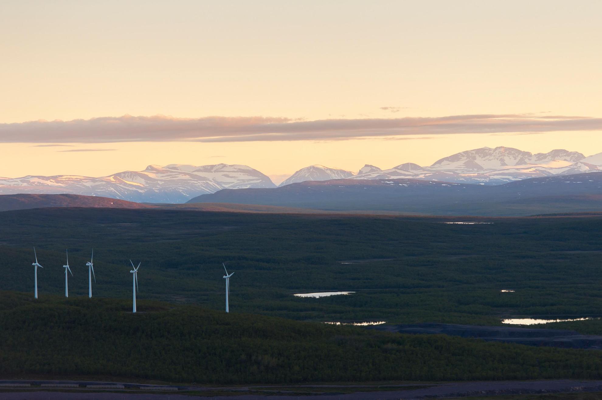 Im Vordergrund eine Tundralandschaft mit fünf Windkraftwerken, im Hintergrund ein Gebirgsmassiv  mit schneebedeckter Bergkette. Der Himmel ist aprikosenfarben.