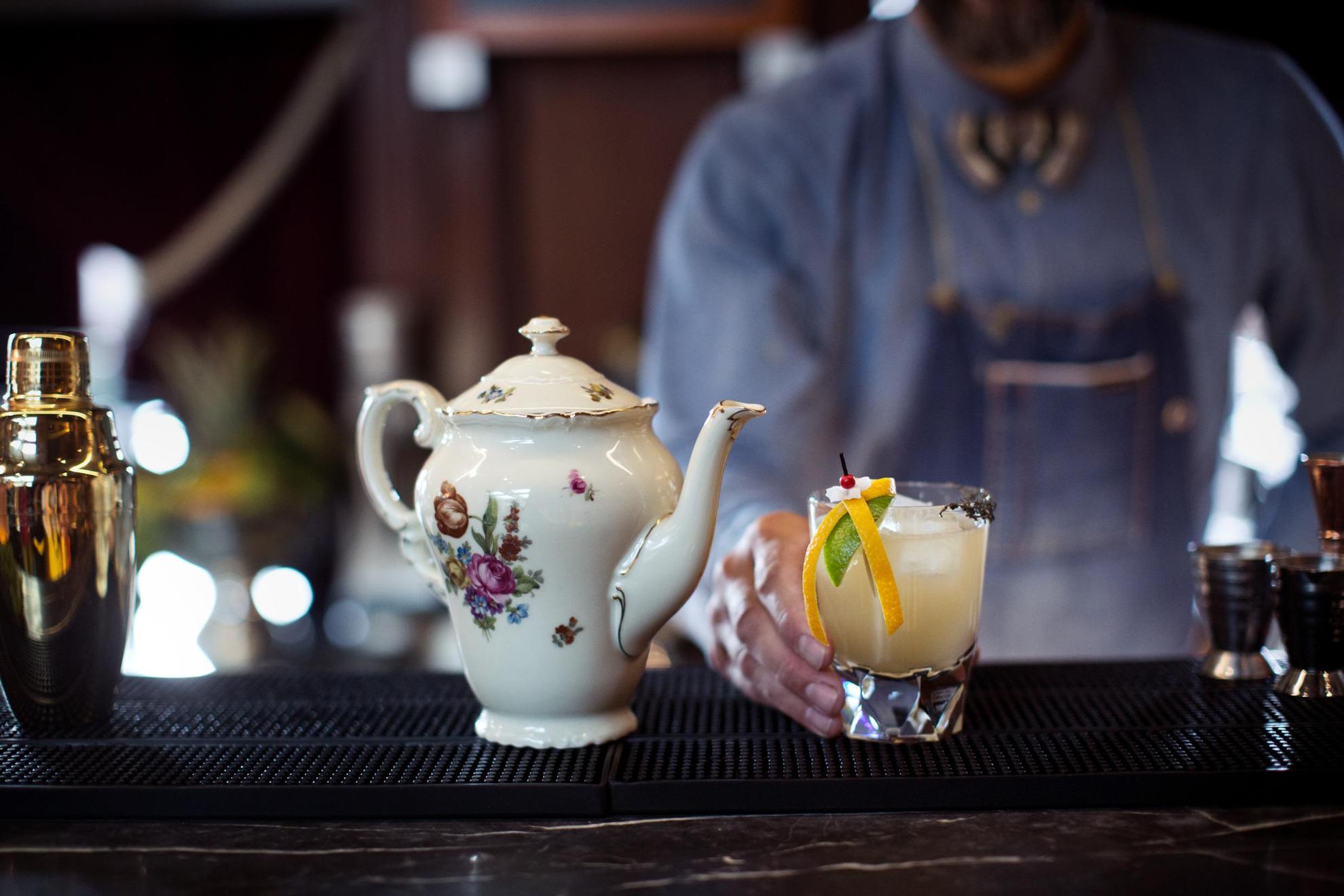 A bartender serving a cocktail decorated with citrus fruit, with a porcelain teapot and a shaker standing next to it.