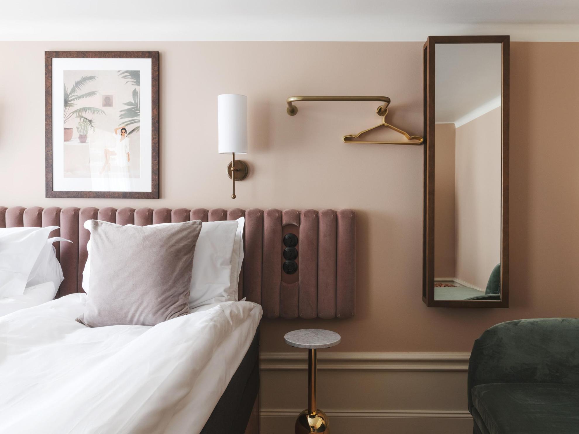 Ein Hotelzimmer mit beige gestrichenen Wänden und einer weißen Decke. Eine Wandlampe aus Messing, ein Gemälde, ein Spiegel und ein Kleiderbügel zieren die Wände neben dem Doppelbett.