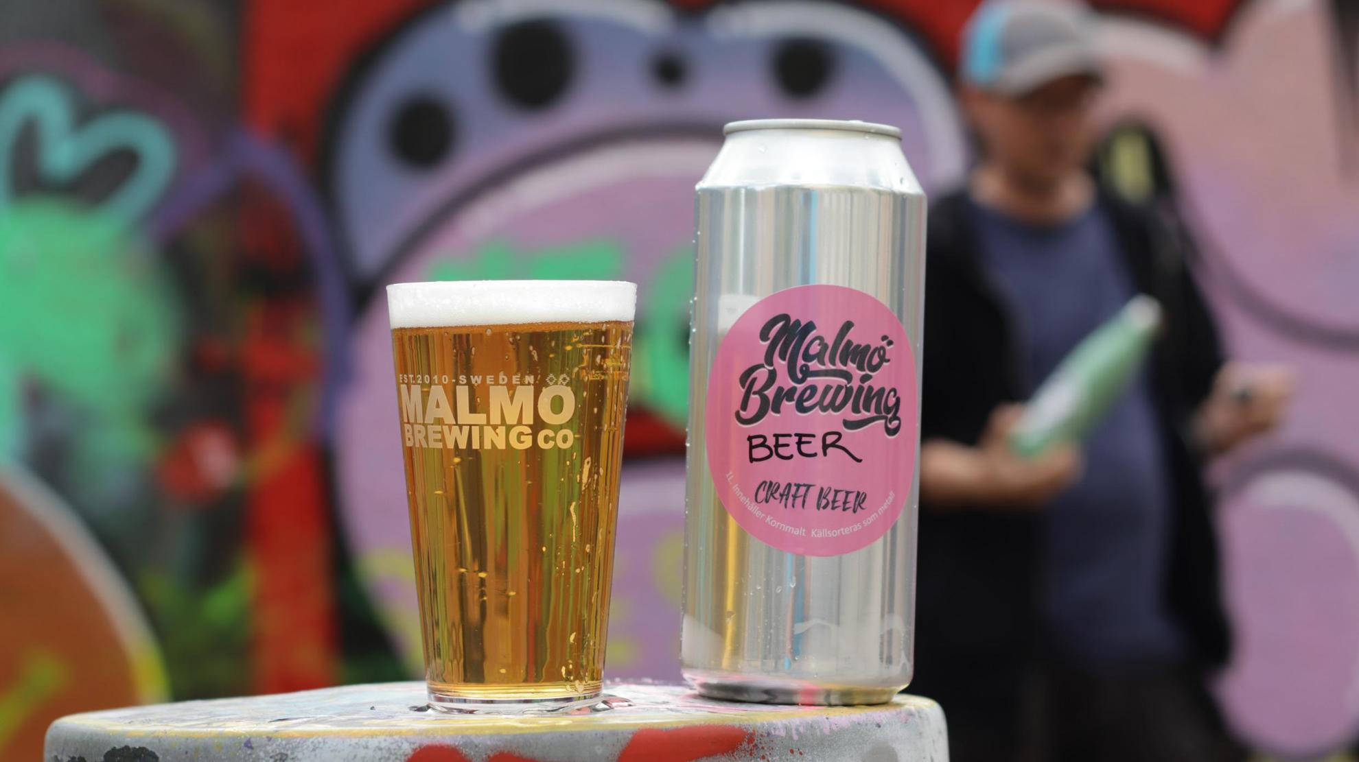 Eine Dose Bier und ein mit Bier gefülltes Glas. Unscharf im Hintergrund ist eine Person und eine Graffitiwand.