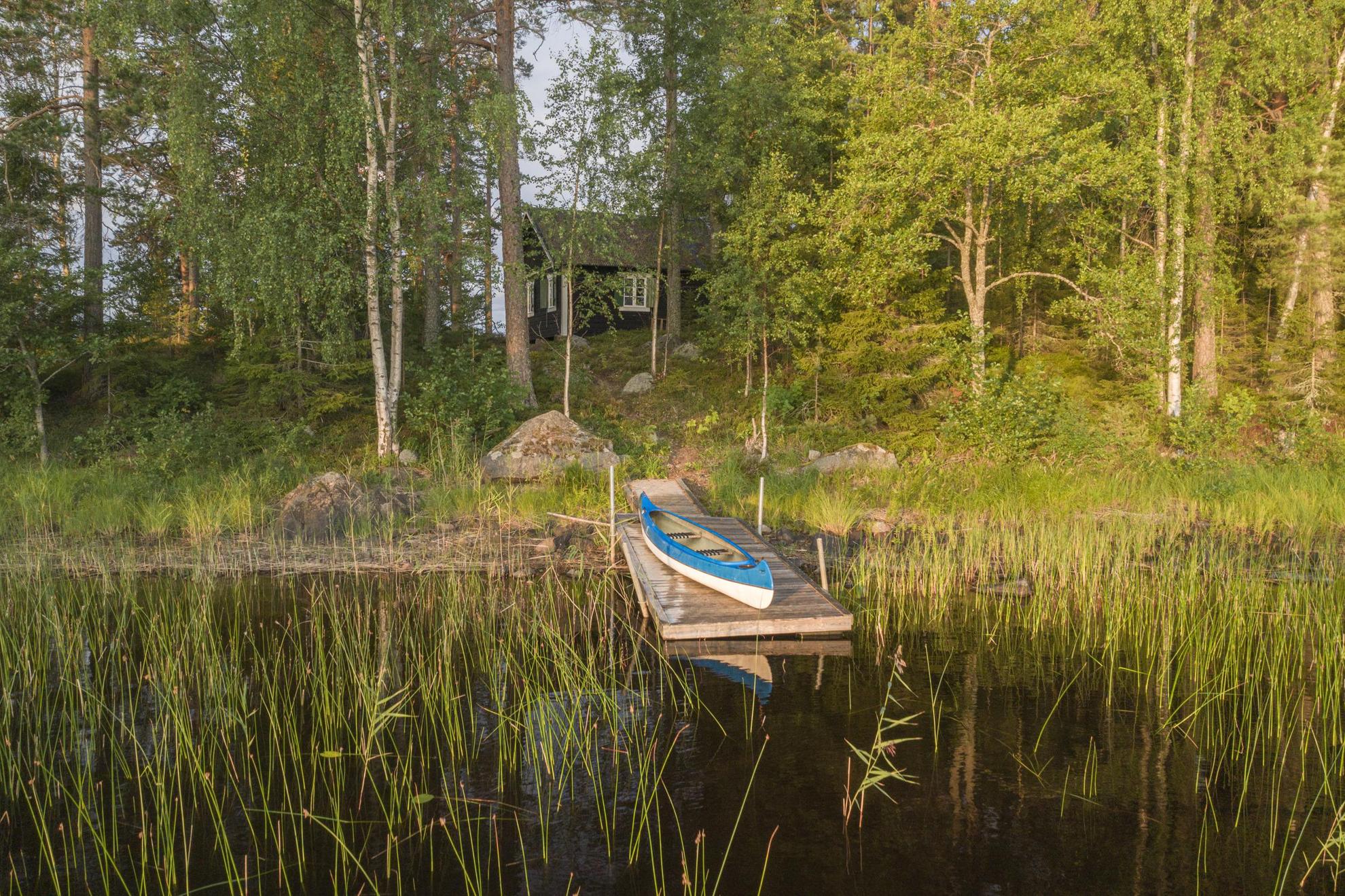 Ein Kanu auf einem kleinen Steg im Schilf eines seichten Sees, wo man eine im Wald versteckte Holzhütte sehen kann.