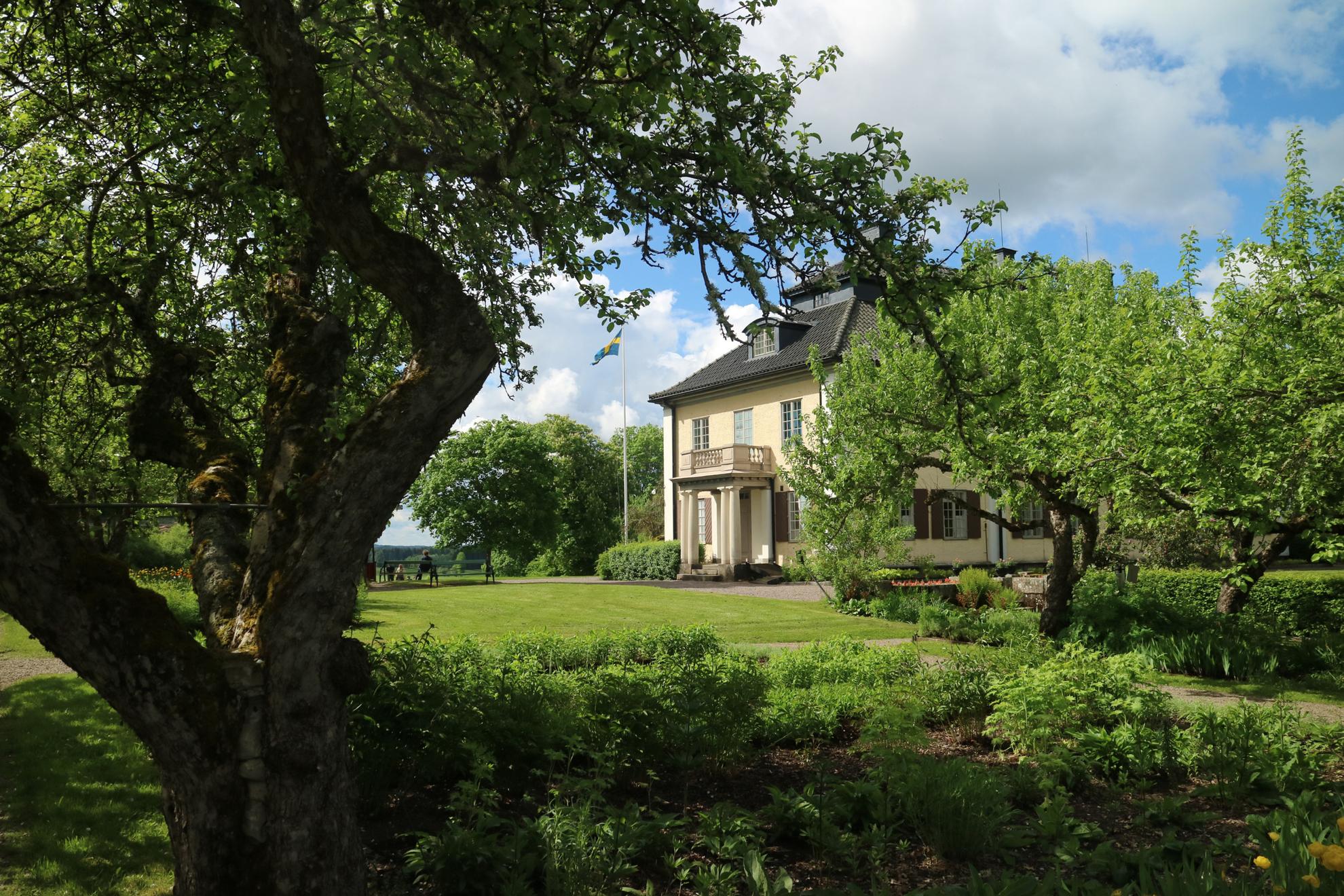 Ein schönes hellgelbes Steinhaus mit einem Garten. Vor dem Haus ein grüner Rasen und rund um das Haus gibt es Bäume, Sträucher und Blumenbeete.