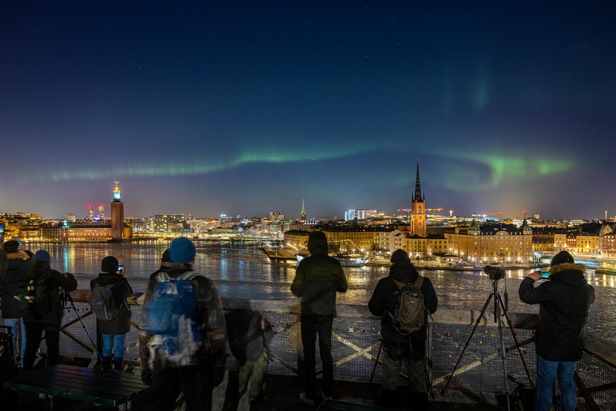 Nordlichter über einer Stadtlandschaft mit Fotografen, die aufgereiht stehen, um ein Foto zu machen.