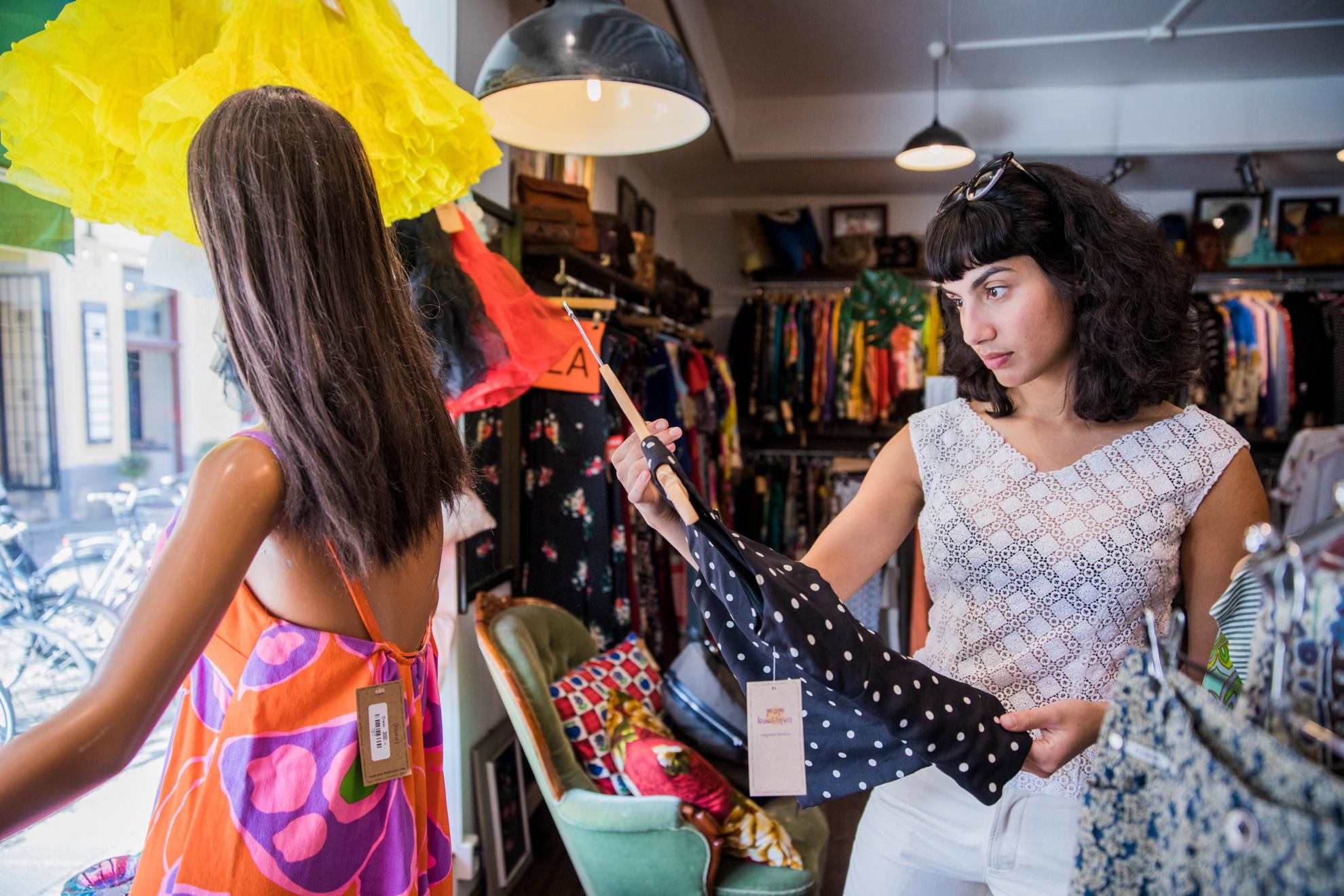 Eine Frau sieht sich eine Bluse in einem Vintage-Laden an. Im Schaufenster neben ihr steht eine Schaufensterpuppe und hinter ihr sieht man viele verschiedene Kleider.