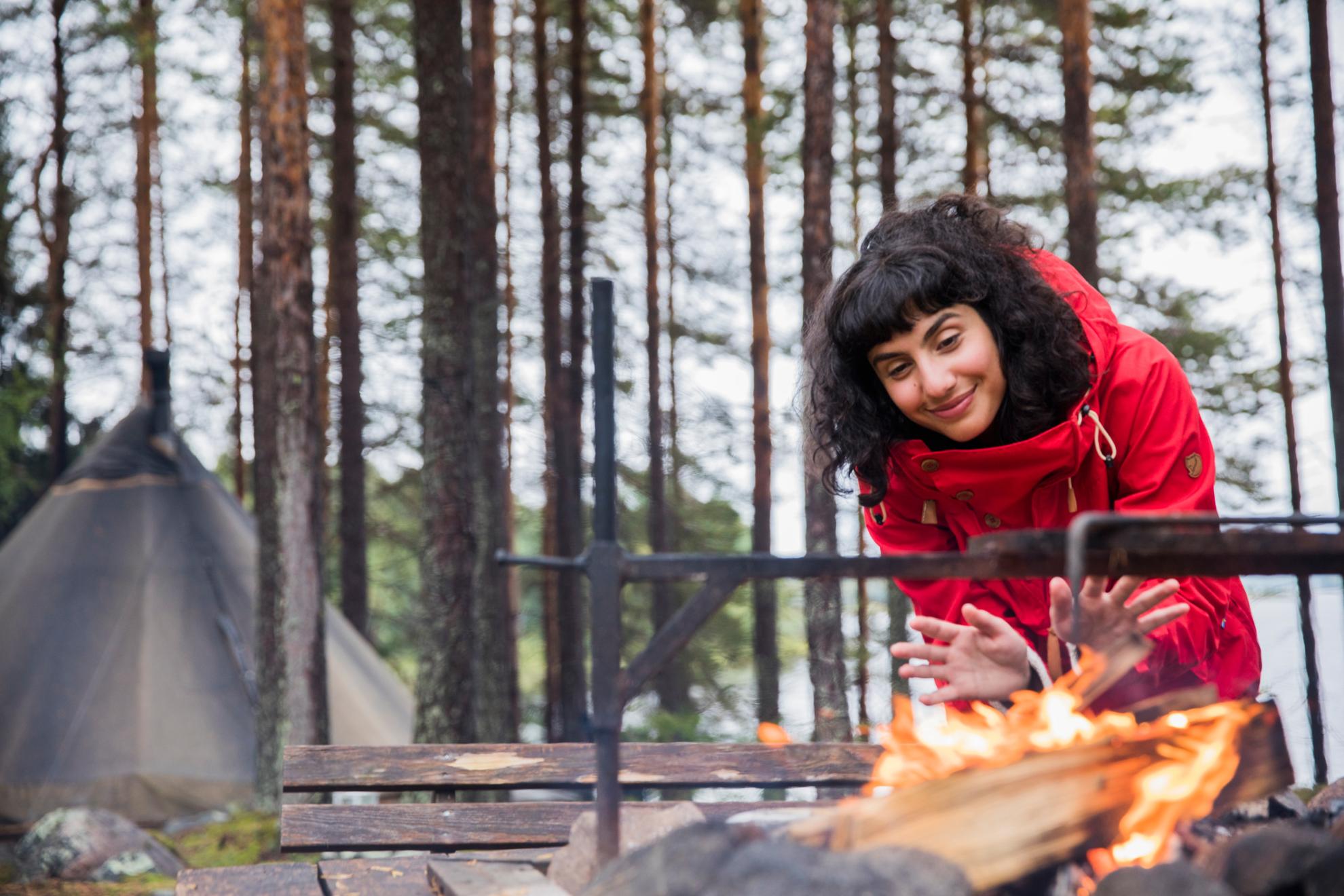 Eine Frau wärmt ihre Hände an einem Lagerfeuer auf einem Glamping-Platz. Hinter ihr ragt ein Zelt auf.