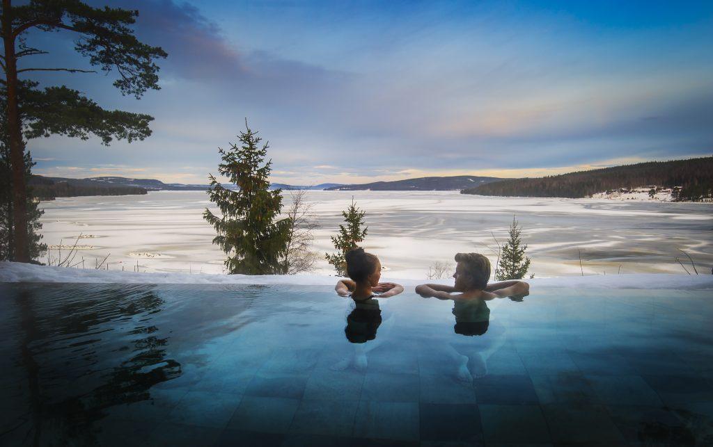 Zwei Personen entspannen sich im Außenpool des Orbaden Spa & Resort in Hälsingland und beobachten den Blick auf das schneebedeckte Wasser.