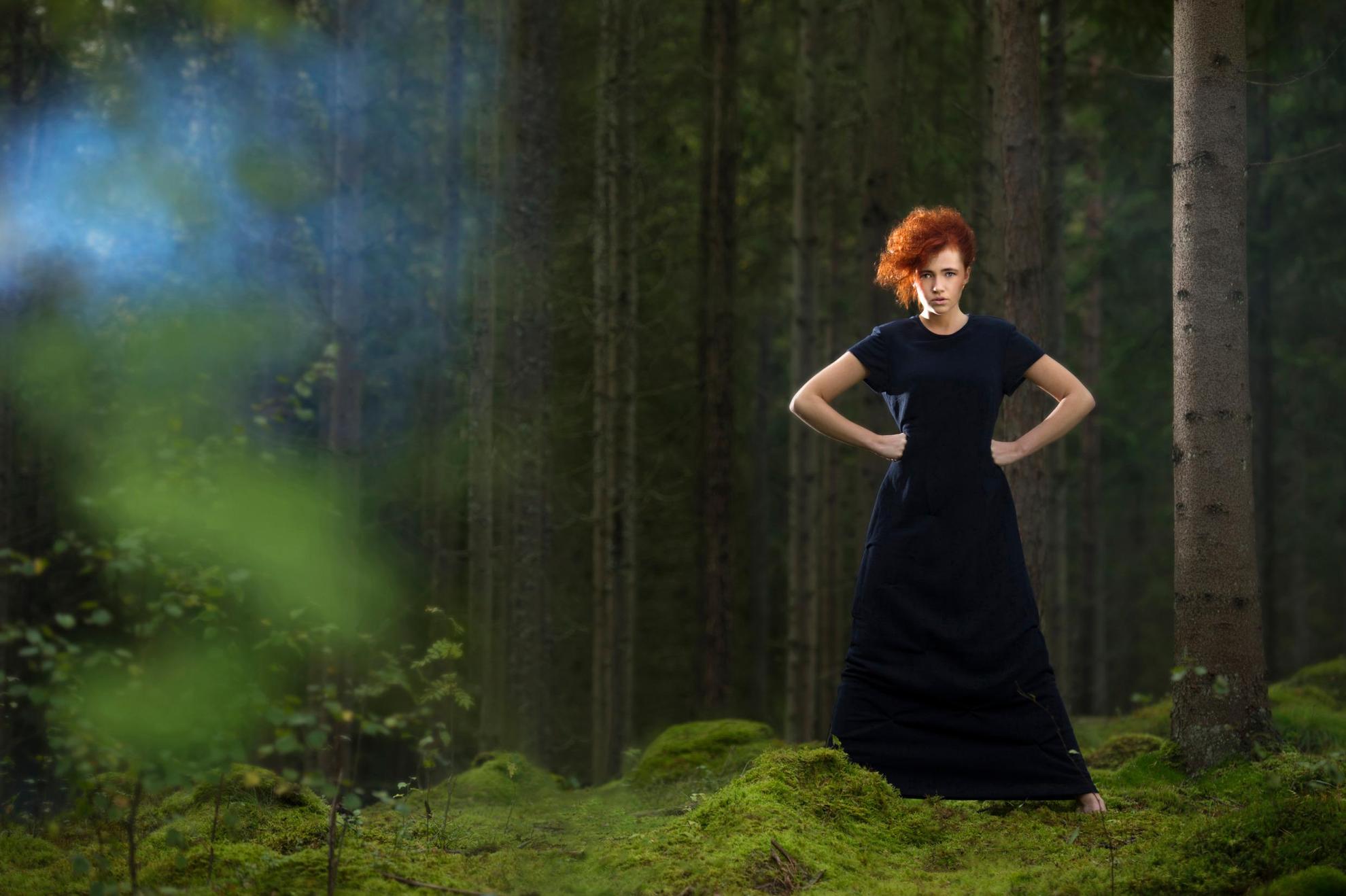 Eine rothaarige Frau trägt ein dunkles Kleid und steht im Wald.