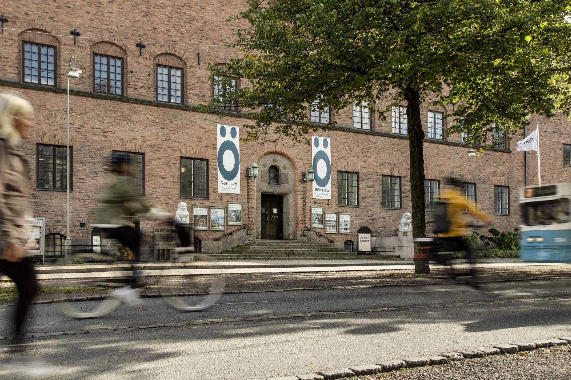 Zwei Radfahrer und eine gehende Person sind verschwommen am Eingang und der Fassade des Röhsska-Museums in Göteborg zu sehen. Das Gebäude ist aus Ziegeln gebaut und auf jeder Seite des Eingangs befinden sich weiße Steinstatuen mit Figuren, die ein bisschen wie Löwen aussehen.