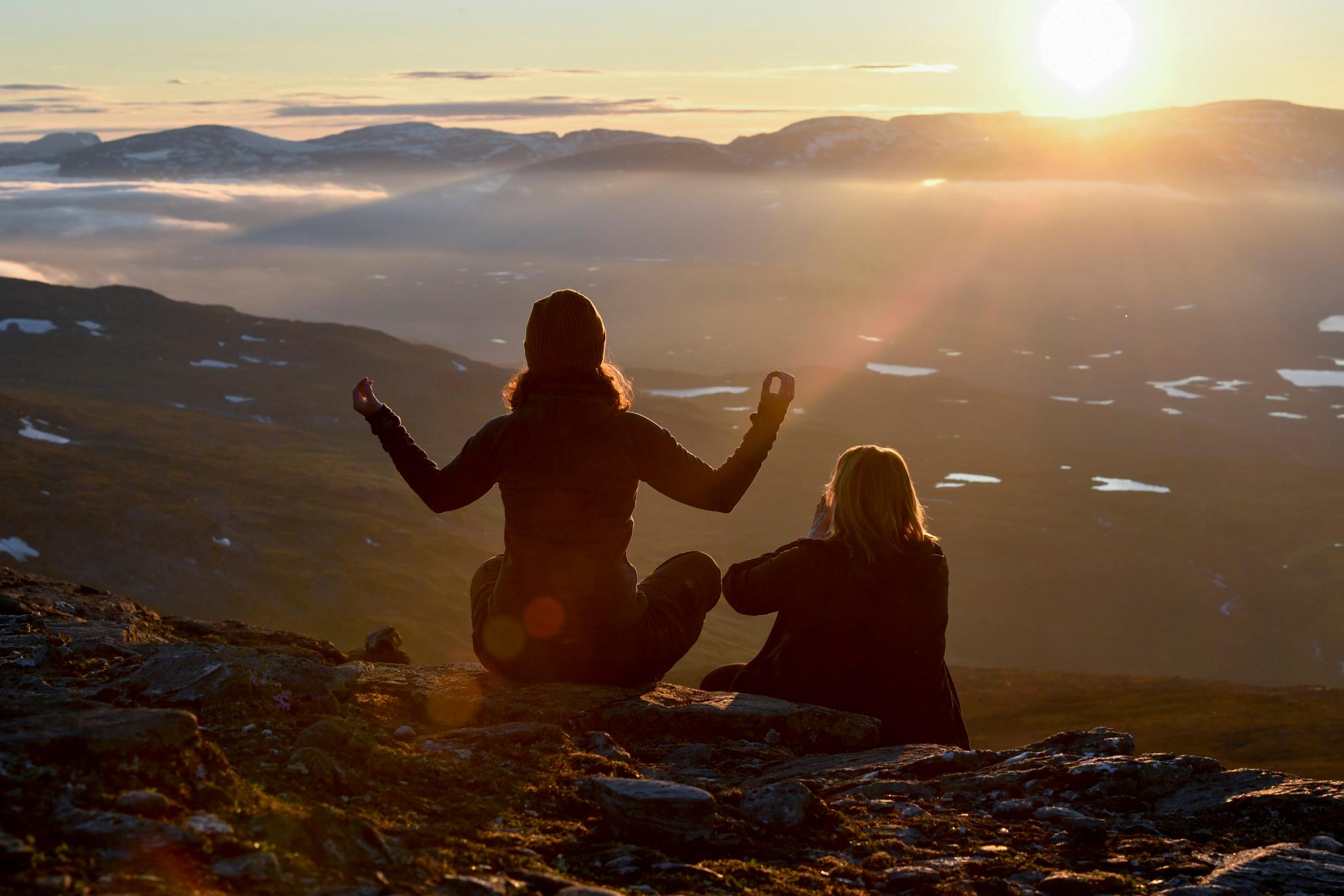 Eine Frau sitzt im Lotussitz neben einer anderen Frau, die ihre Handflächen aneinander presst, vor Bergen und einem Sonnenuntergang.