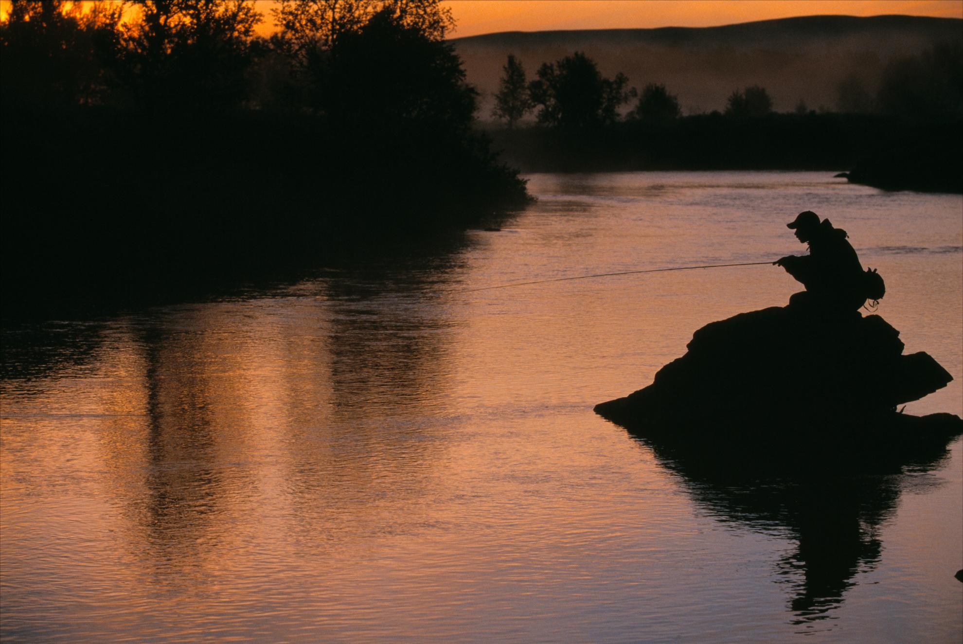 Es ist Nacht und eine Person kauert auf einem Felsen in einem Fluss bei Ammarnäs und hält eine Angel in der Hand.