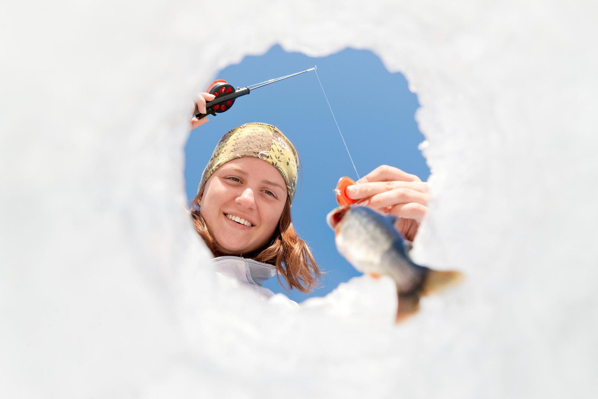 Eine Person schaut durch ein Eisloch und hält ein Fisch an der Angelleine.