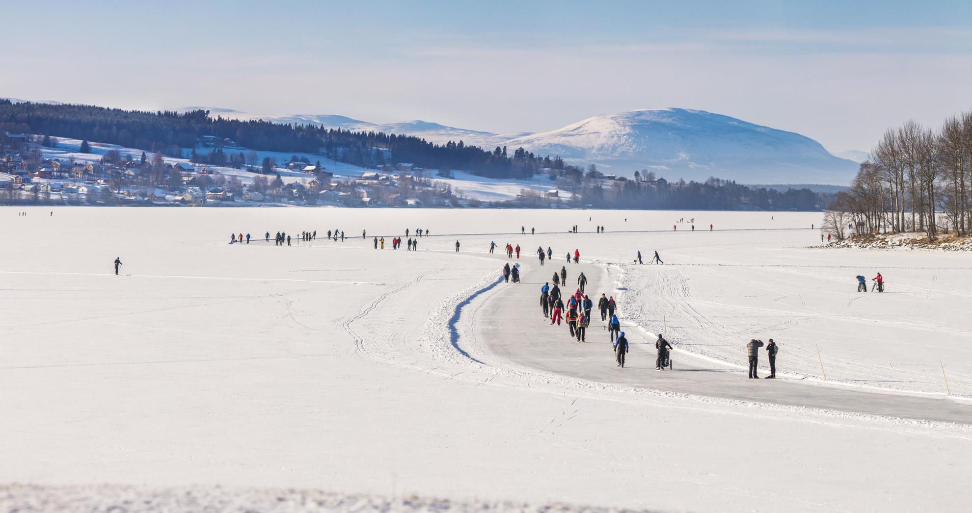 Auf einem schneebedeckten, zugefrorenen See laufen mehrere Personen auf präparierten Bahnen Schlittschuh. Häuser, Bäume und Berge im Hintergrund.