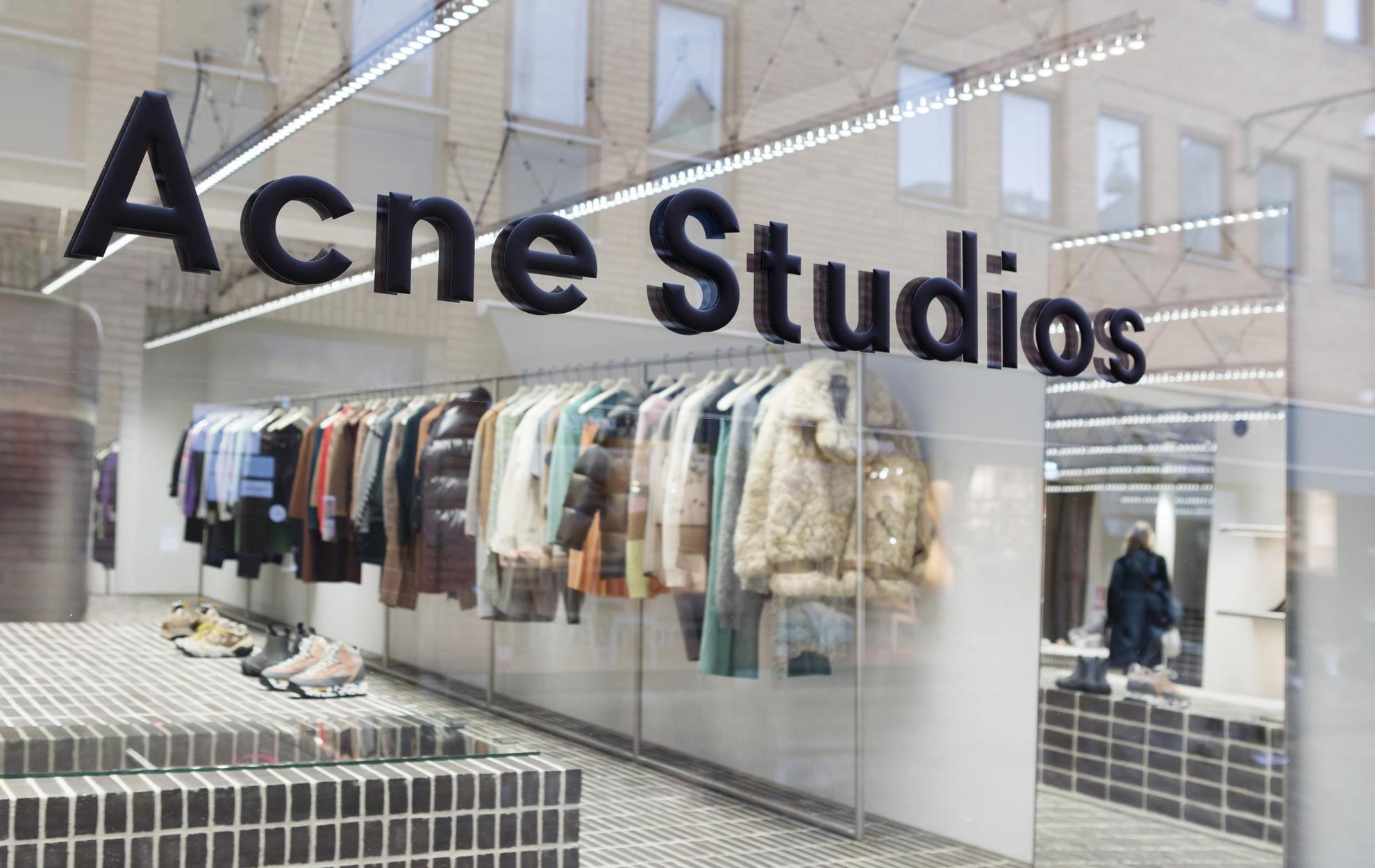 Das Schaufenster des Acne Studios in Stockholm. Auf einer  Kleiderstange entlang der Wand hängen zahlreiche Kleidungsstücke.