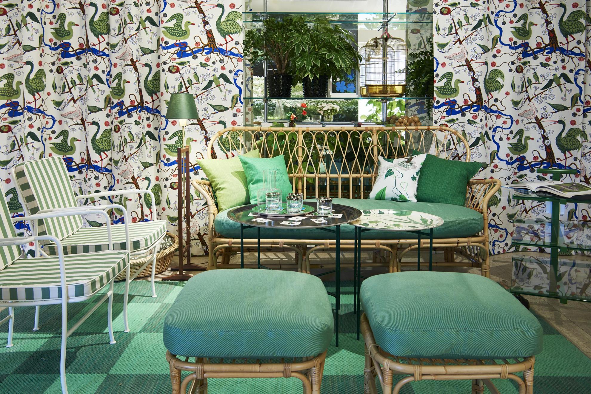 Eine grüne Ecke in einem Designgeschäft. Ein Sofa, zwei Sessel und zwei Hocker um einen Tisch herum. Hinter der Möbelgruppe befinden sich bunte Vorhänge mit Vögeln in Grün.