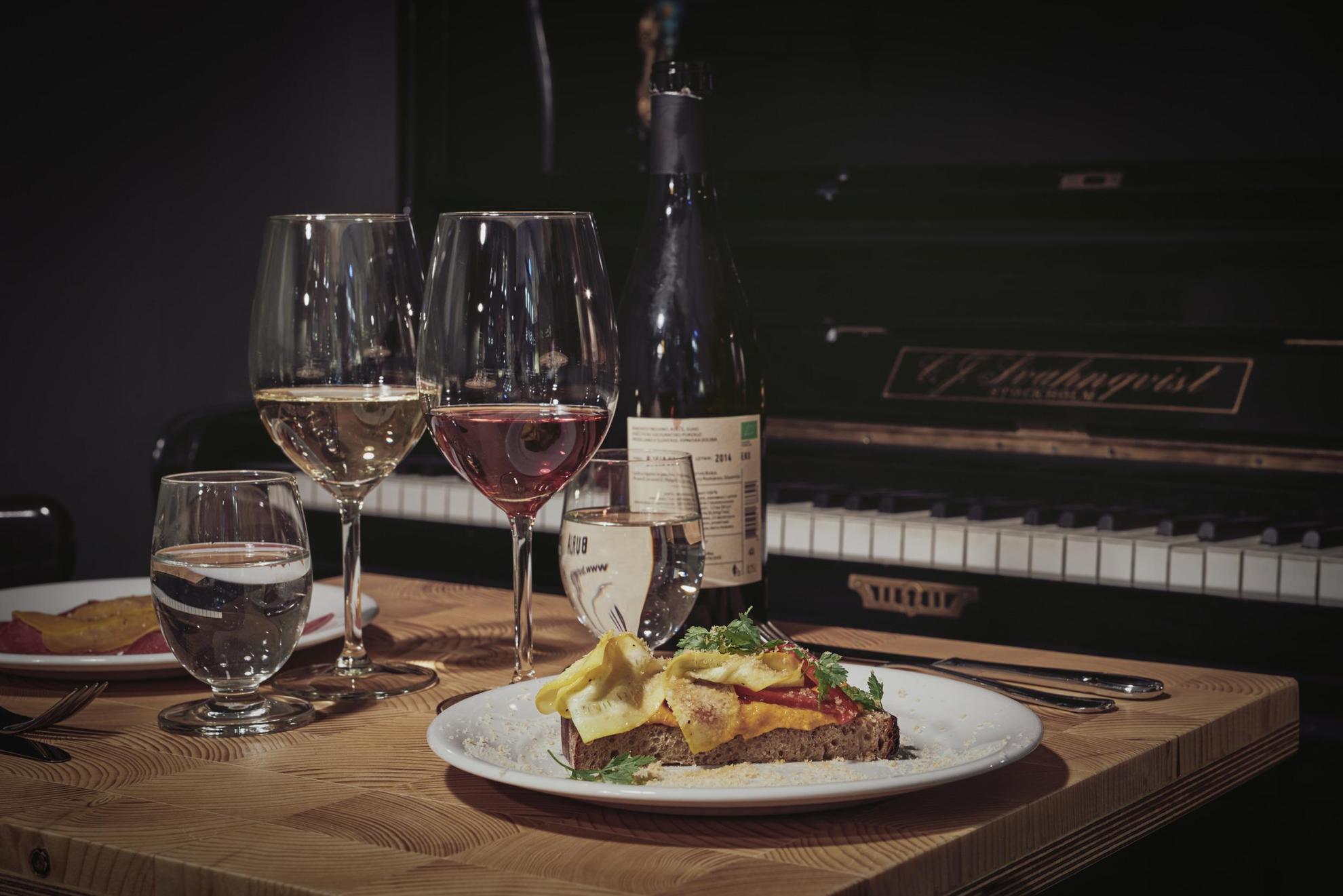 Ein gedeckter Tisch mit einer Weinflasche, zwei Tellern mit Essen und zwei Weingläsern. Der Tisch steht neben einem Klavier.