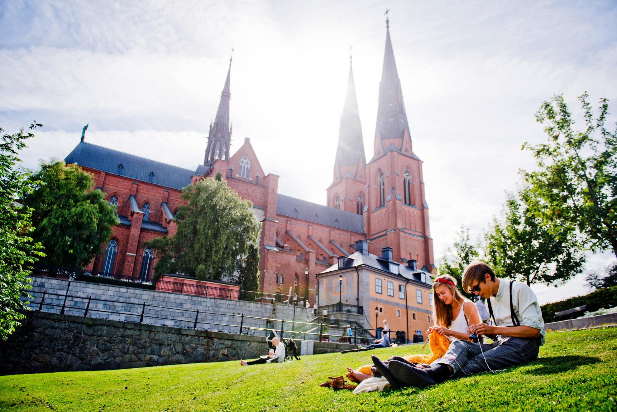 Sonniger Blick auf den Dom von Uppsala mit Menschen im Vordergrund, die auf dem grünen Gras sitzen.
