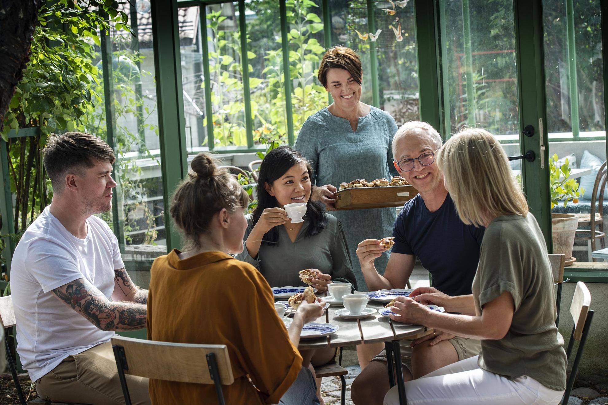 Eine Gruppe von Freunden sitzt an einem Tisch in einem Café und trinkt Kaffee und Zimtschnecken. Neben dem Tisch steht eine Frau mit einem Tablett voller Zimtschnecken.