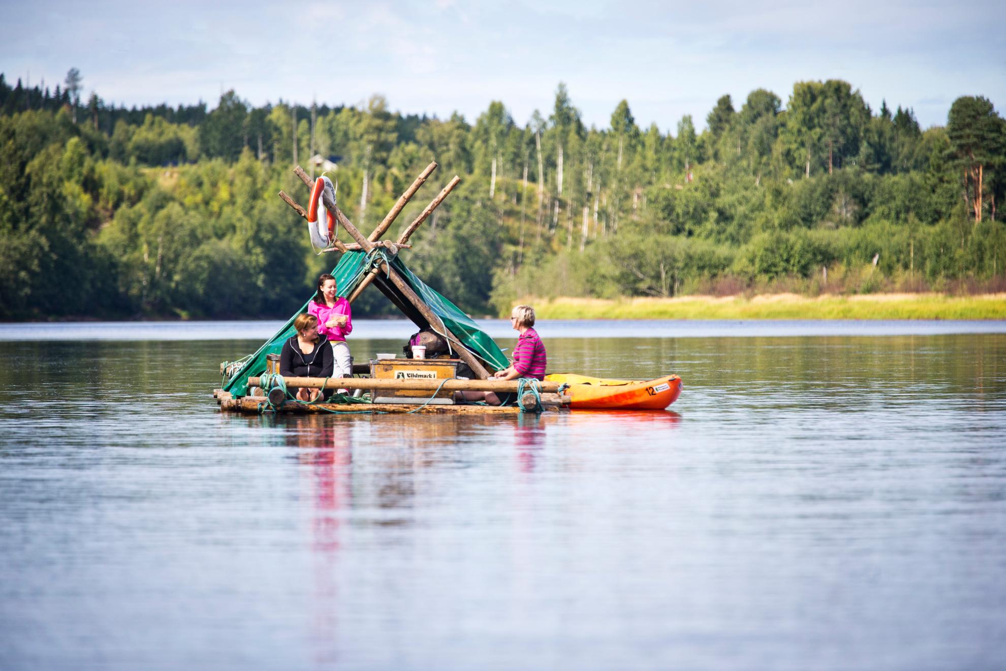 Drei Frauen lassen sich auf einem Holzfloß einen Fluss entlang treiben. Neben dem Floß steht ein Kanu. Der Fluss ist ruhig und im Hintergrund ist ein Wald zu sehen.