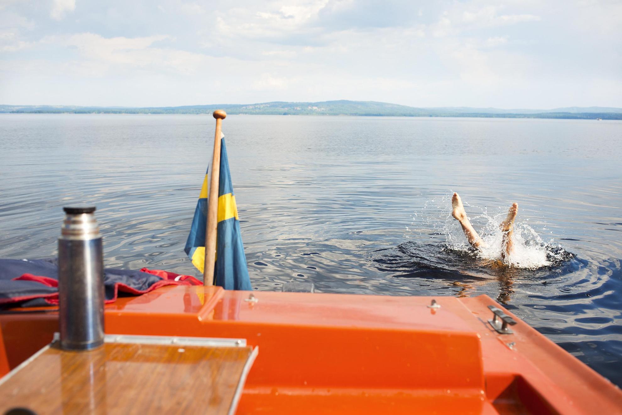 Das Heck eines kleinen orangefarbenen Bootes mit einer schwedischen Flagge.  Eine Person ist gerade aus dem Boot getaucht und man sieht nur die Beine im Wasser.