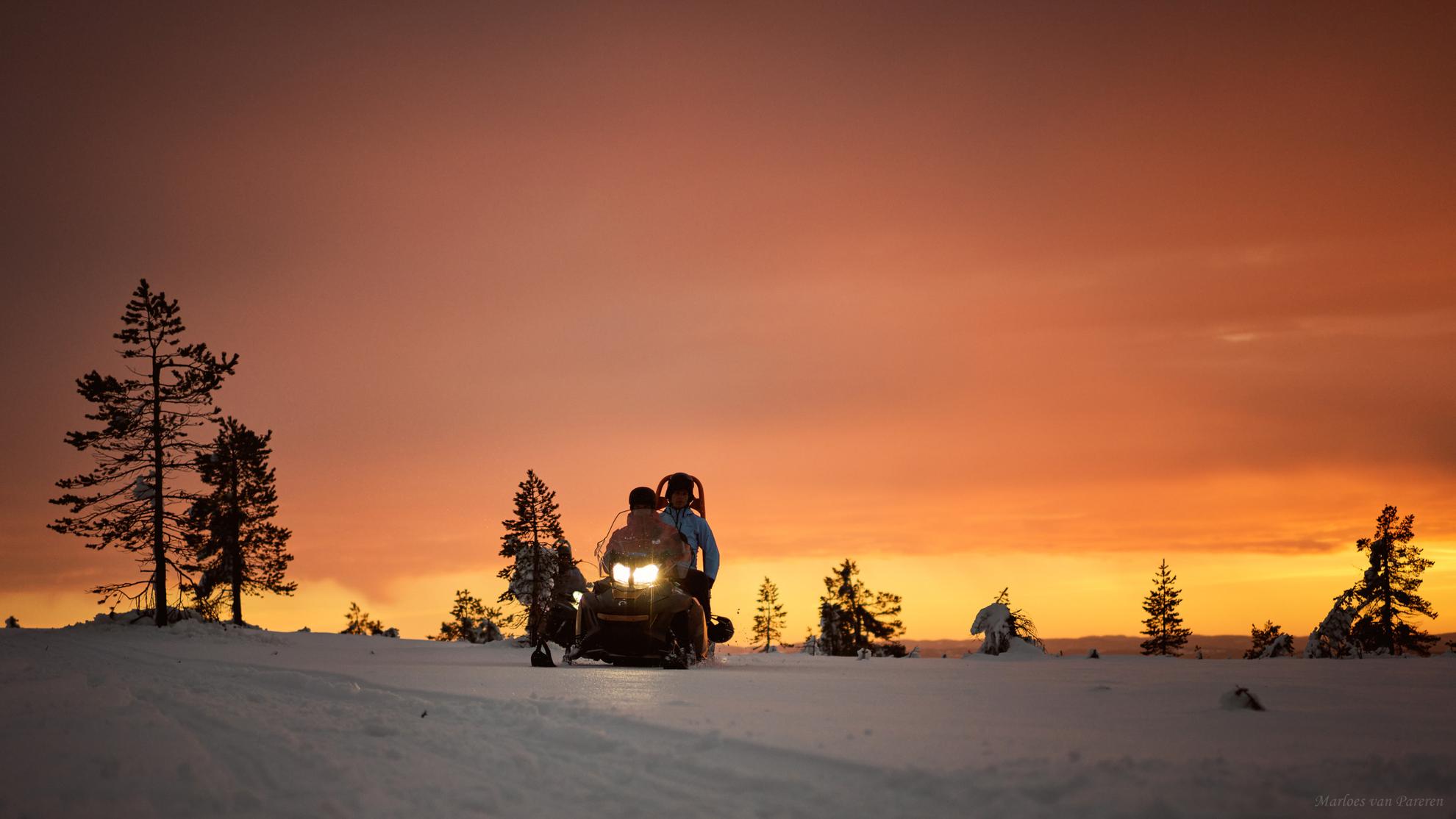 Zwei Personen sitzen auf einem Schneemobil in Schweden. Der Himmel ist durch das orangene Licht der Abendsonne gefärbt.