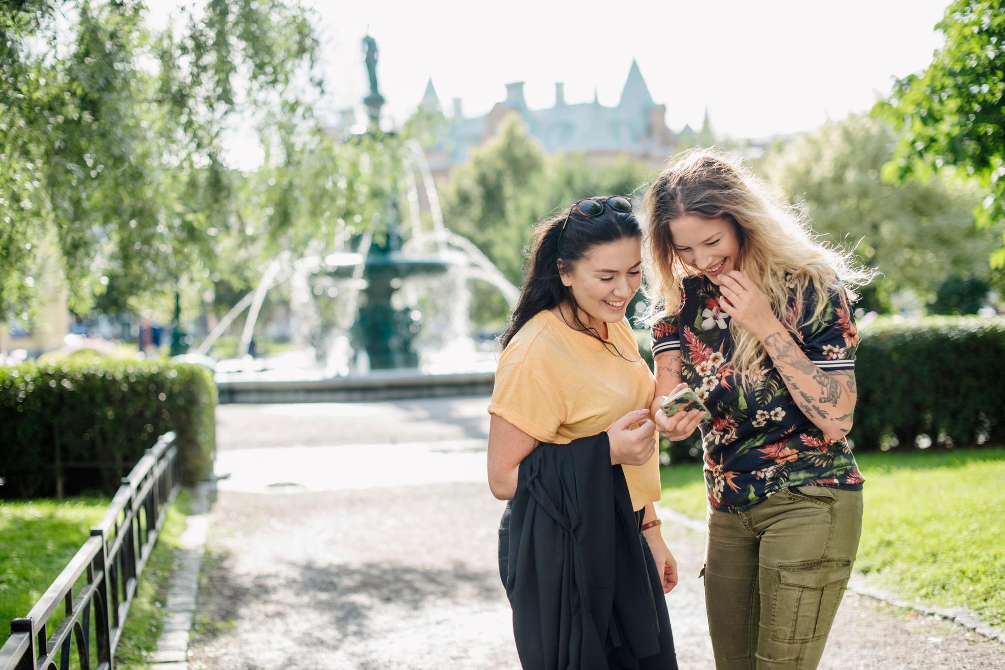 Zwei Frauen, die in einem Park auf ein Mobiltelefon schauen, mit einem Springbrunnen im Hintergrund.