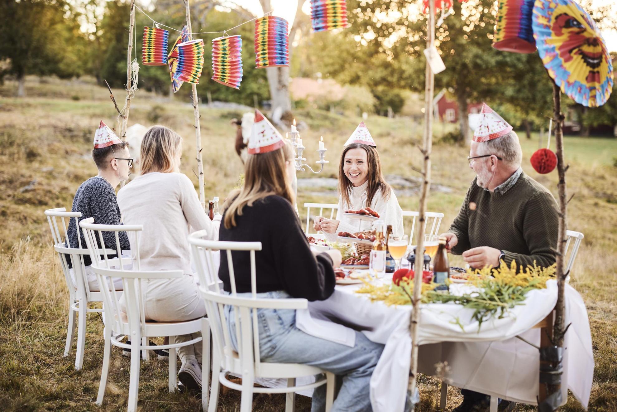 Eine Krebsparty im Freien. Mehrere Personen sitzen um einen Tisch versammelt und genießen Krebse. Sie tragen typische Partyhüte und stoßen mit Gläsern an.