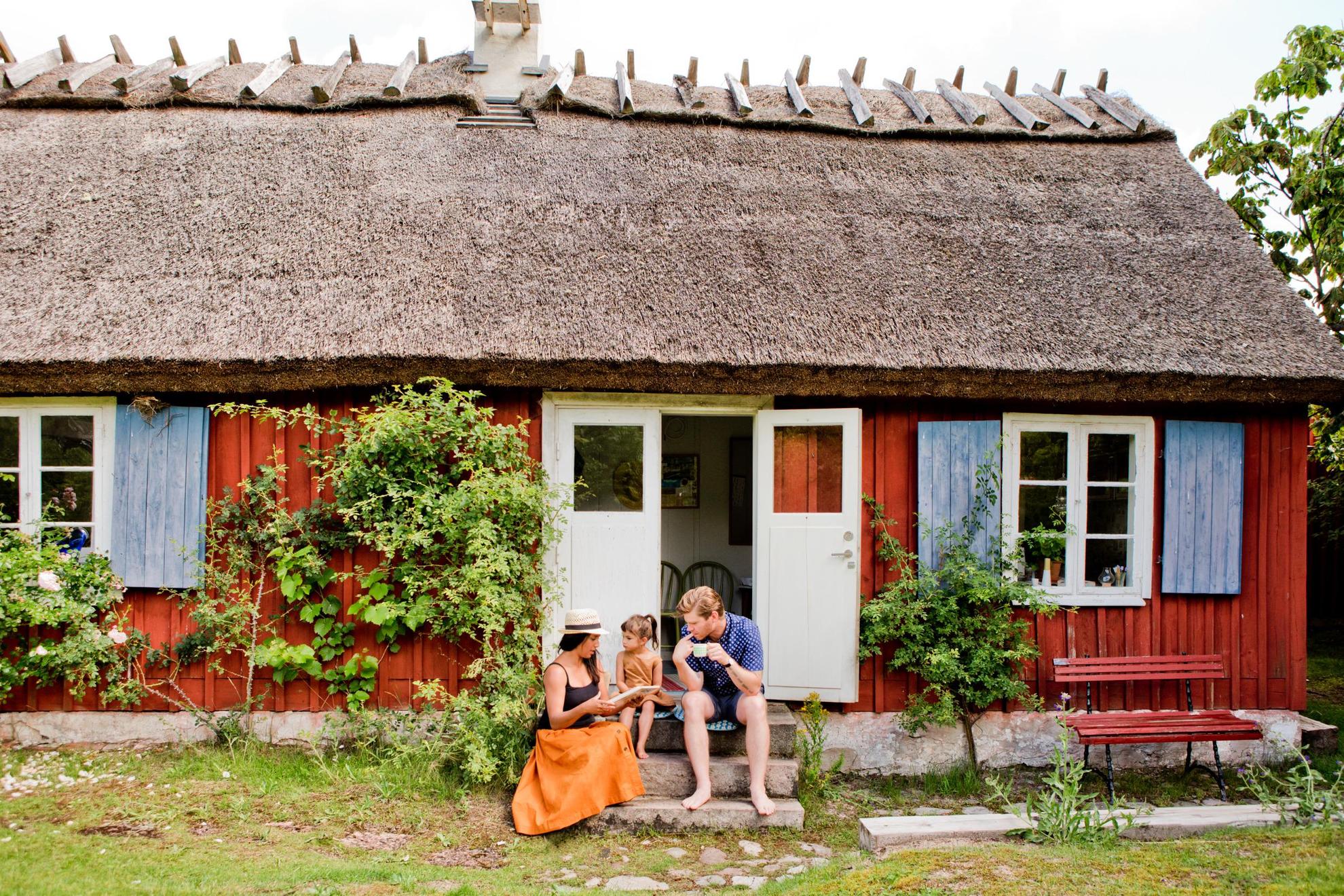 Ein rotes Schwedenhaus mit blauen Fensterrahmen und einer blauen Tür. Eine Familie sitzt auf den Stufen zur Haustür.