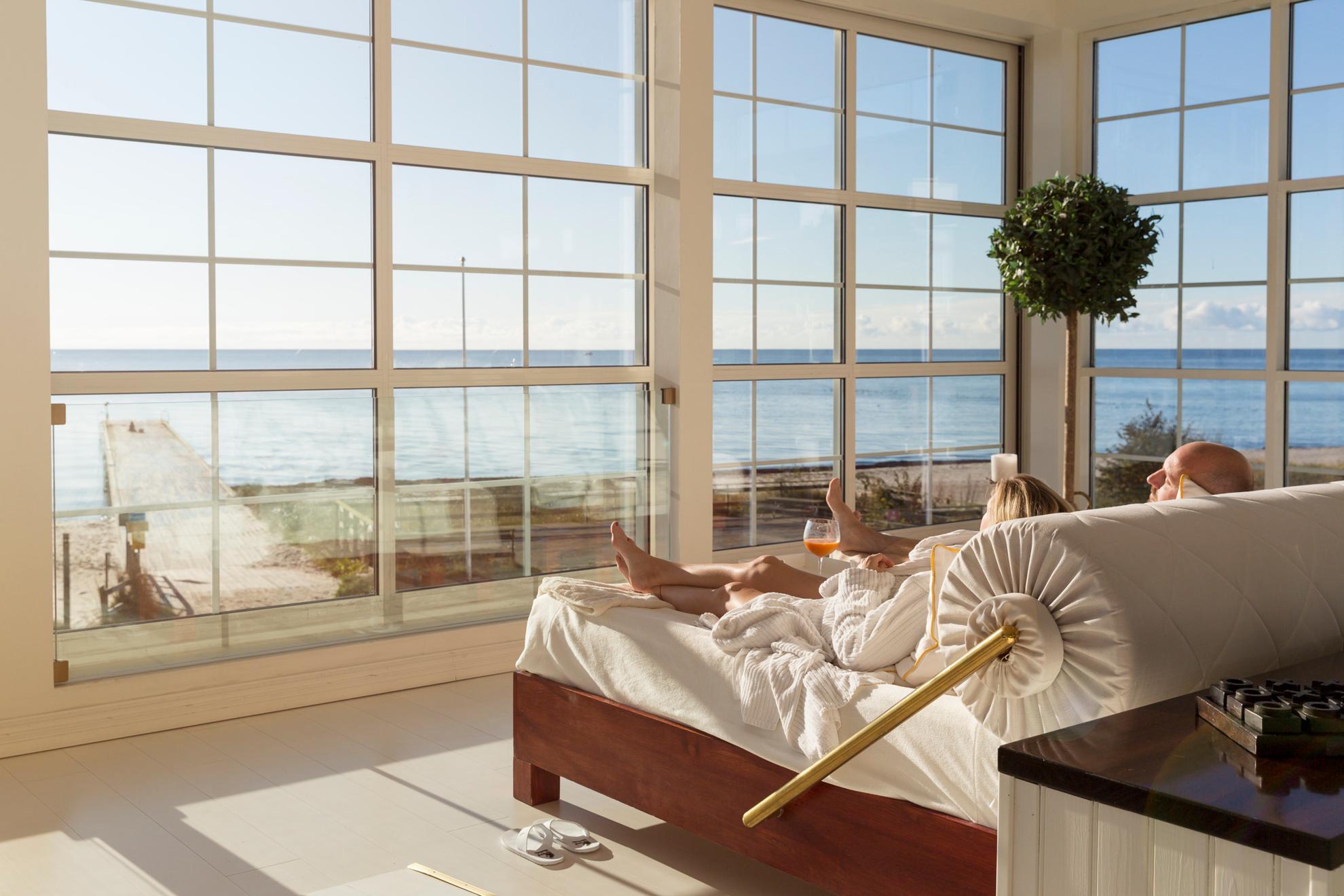 Ein Paar liegt in einem großen Bett neben großen Fenstern mit Blick auf den Strand und das Meer.