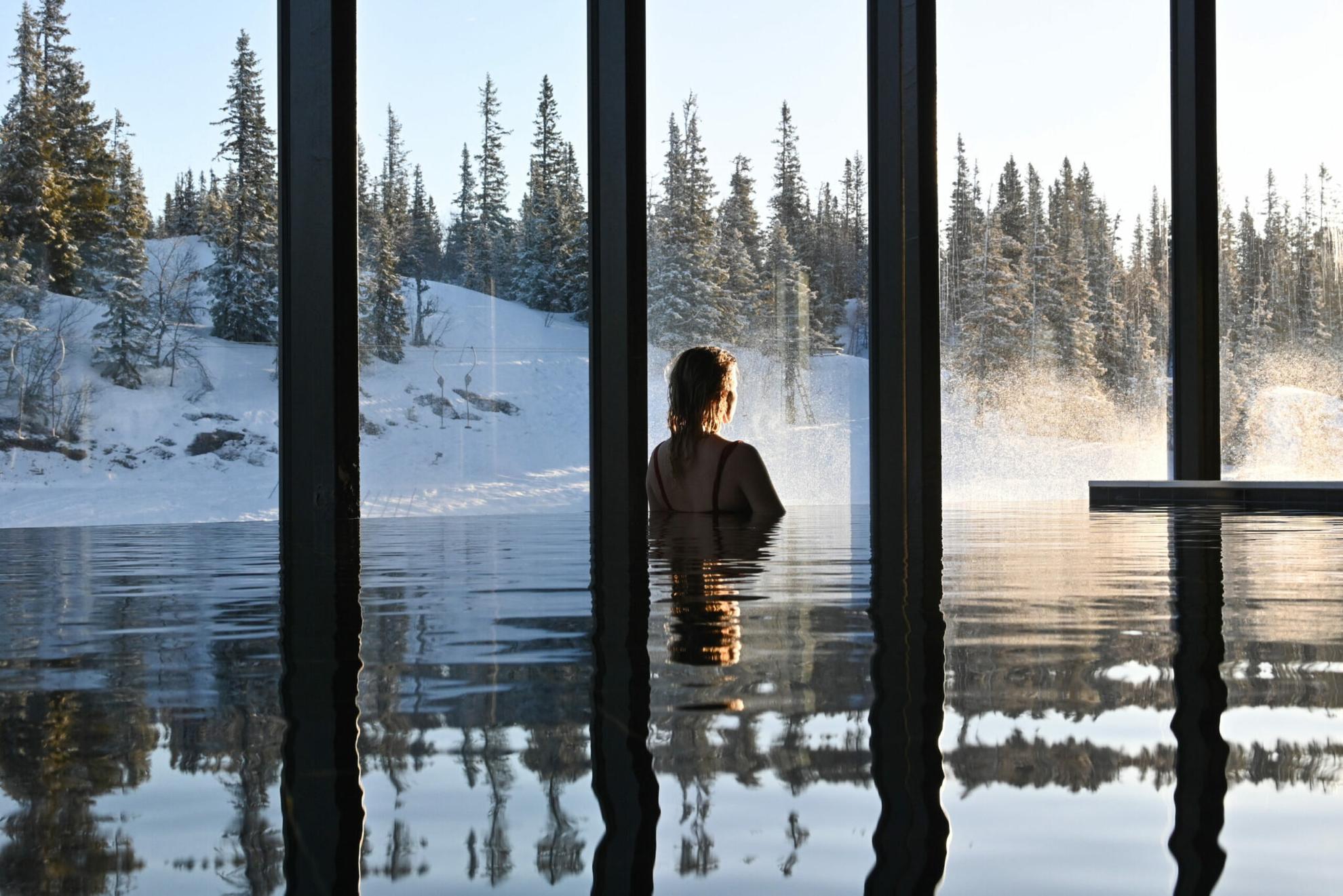 Eine Person in einem Infinity-Pool, die durch die großen Fenster auf den schneebedeckten Wald blickt.