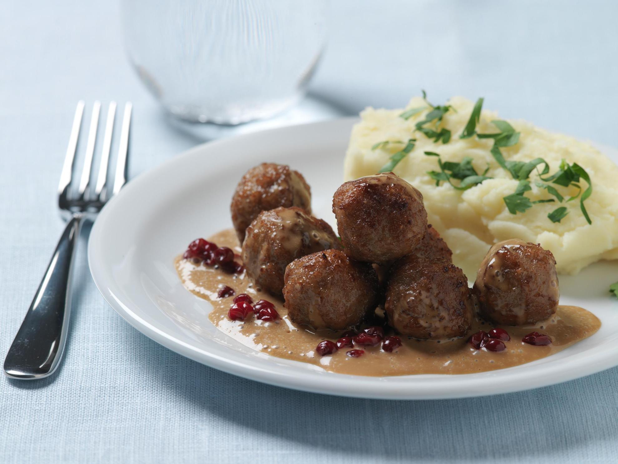 A table is set with a plate of meatballs, mashed potatoes, brown gravy and lingonberry jam next to a glass and fork.