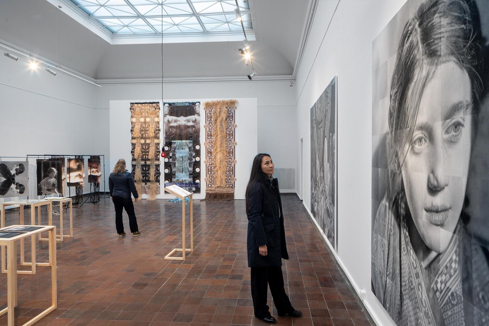 Eine Installation in einer Ausstellung. Eine Frau betrachtet ein großes Schwarz-Weiß-Bild einer Frau und im Hintergrund betrachtet eine weitere Frau ein großes Kunstwerk mit Fotografien, die mit Fragmenten zu neuen Strukturen verflochten sind.