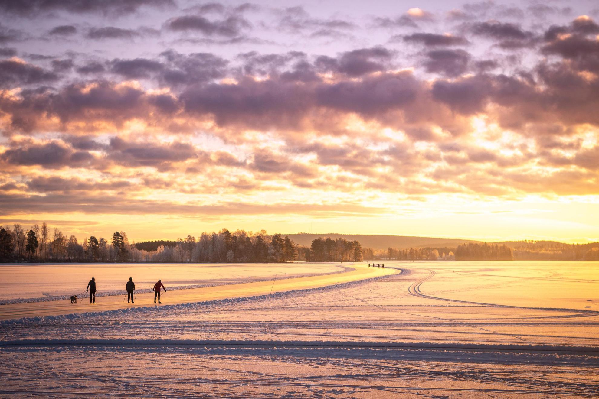 Die Silhouette von drei Personen beim Schlittschuhlaufen auf einem zugefrorenen See. Die Sonne leuchtet orange.